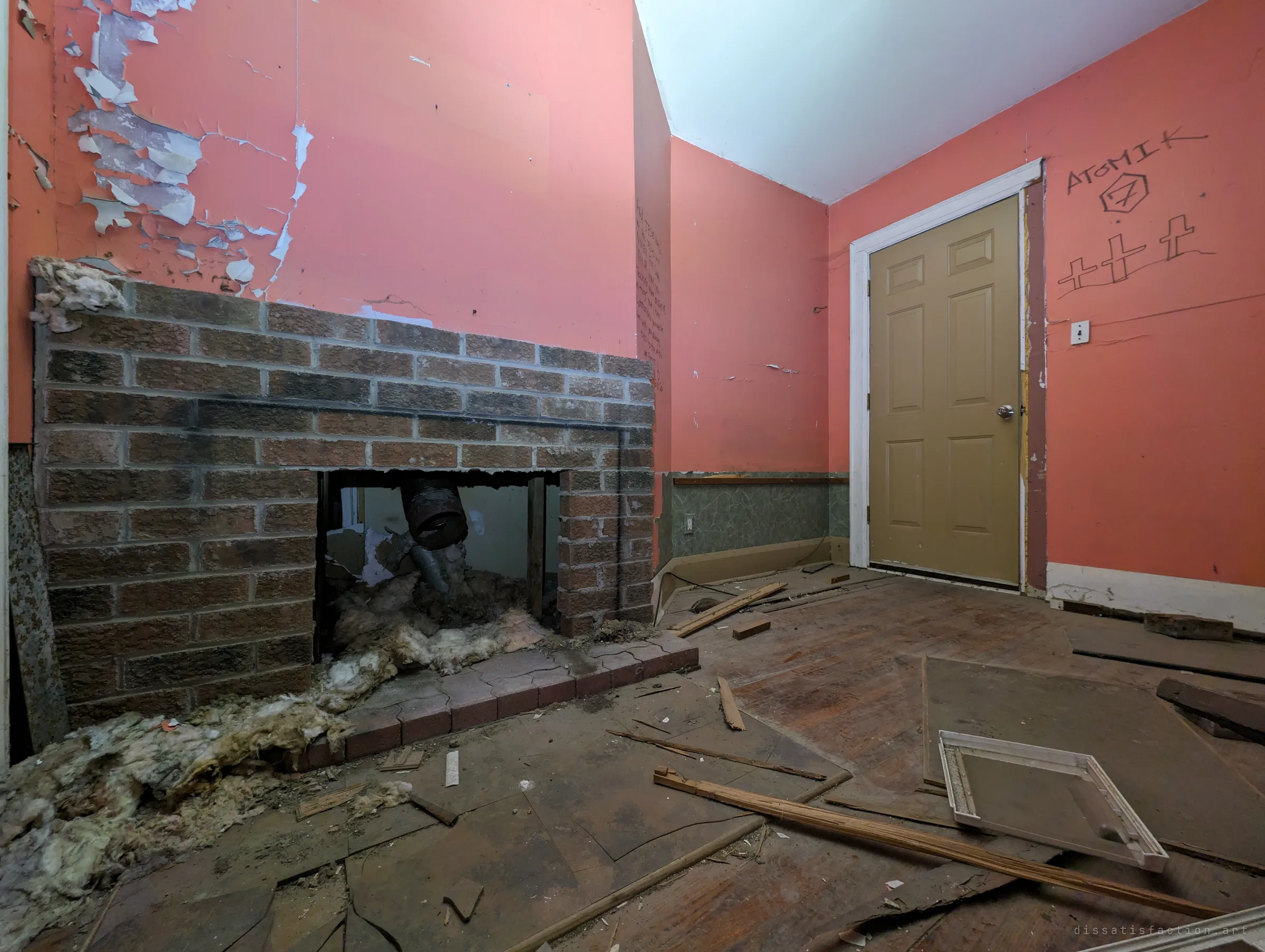 A coral-pink room with a brick fireplace gutted down to its flue pipe and insulation spilling onto the floor. A beige door stands closed beside marker drawings of crosses.