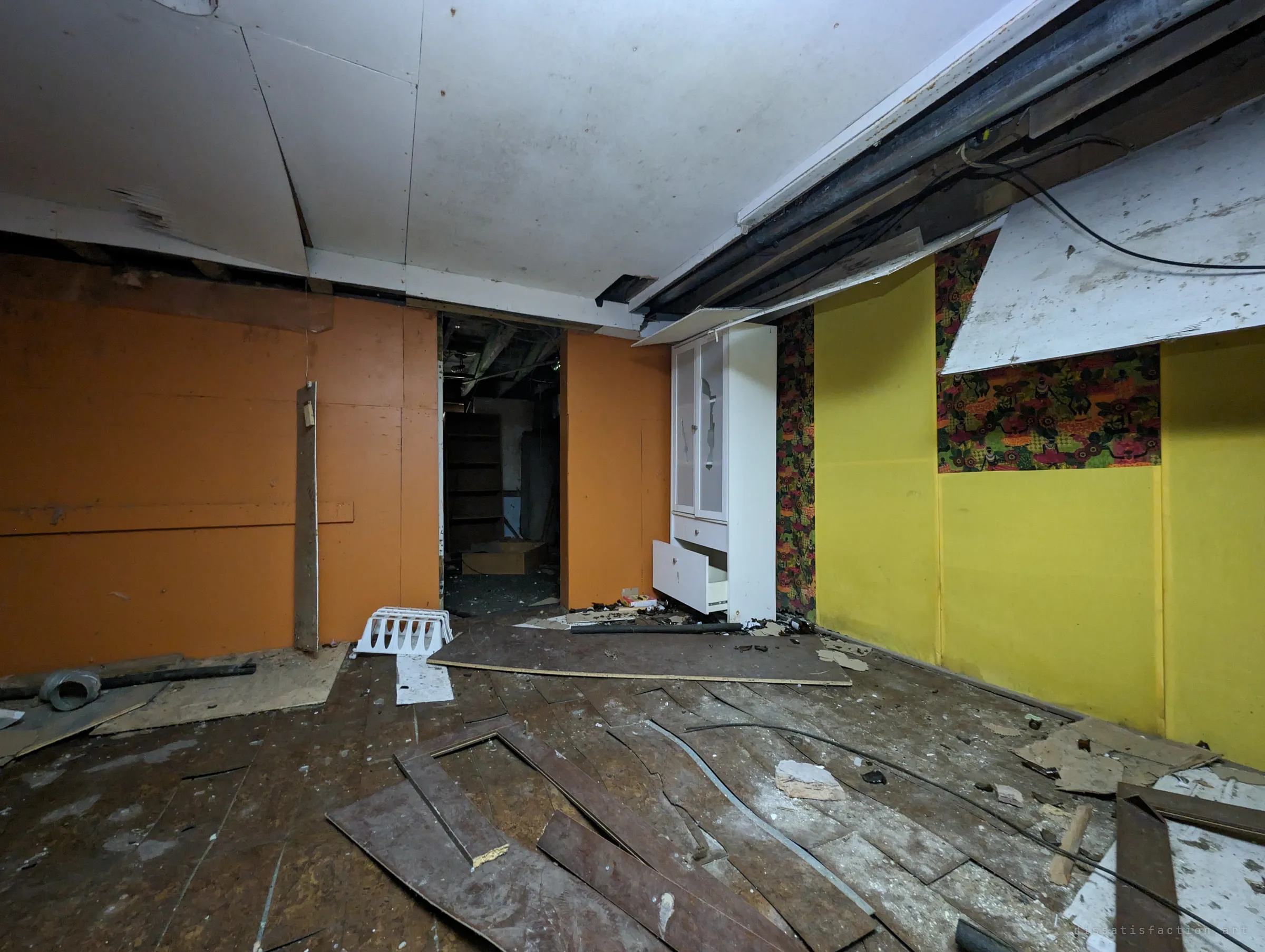 A basement room with orange and yellow panelled walls, a strip of loud floral wallpaper and a warped wooden floor. A doorway leads into a darker storage room.