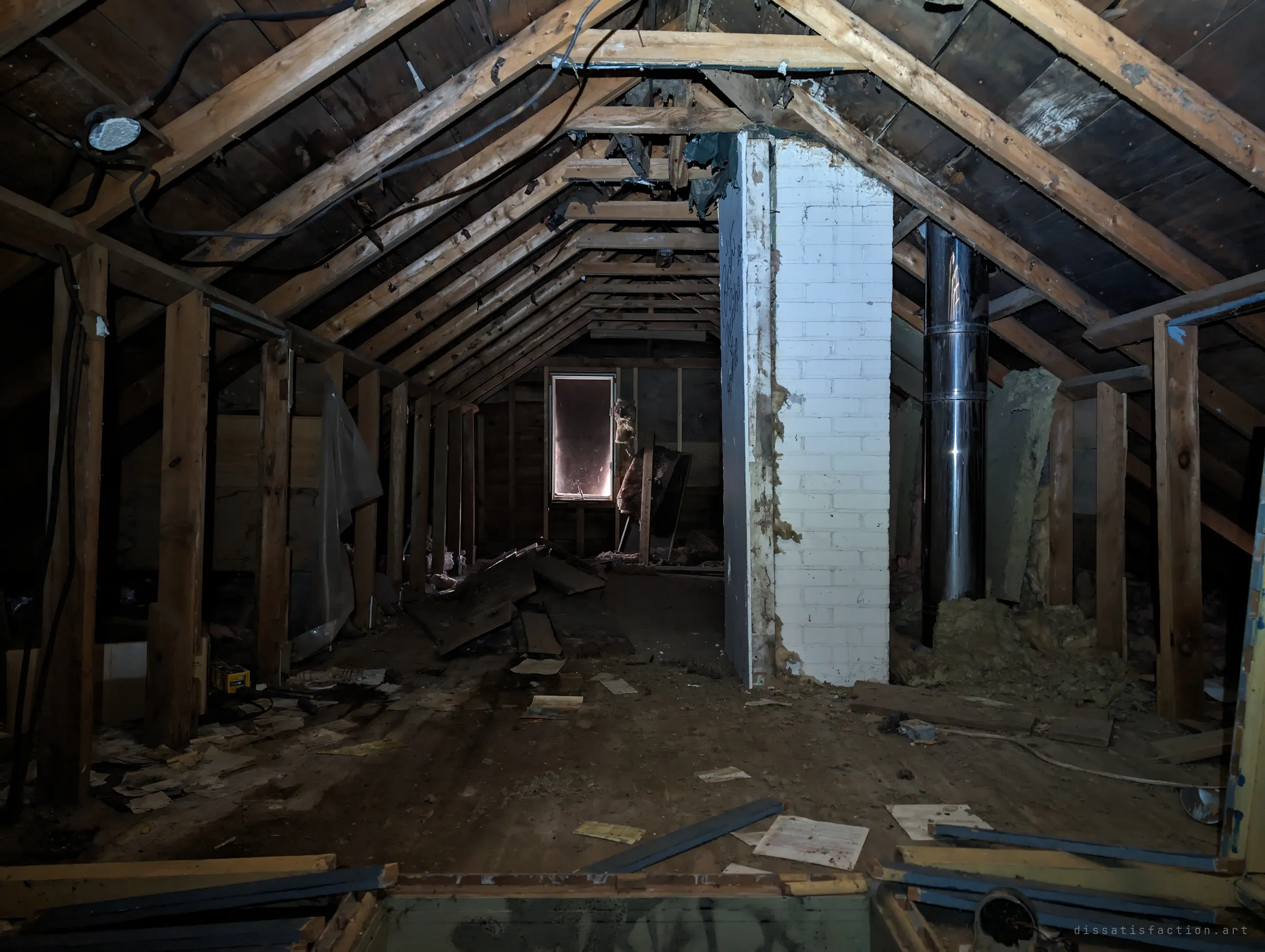 The attic under bare wooden rafters, with a white brick chimney and a steel flue running up the middle. Old papers and boards lie scattered across the dusty floor toward a small lit window.