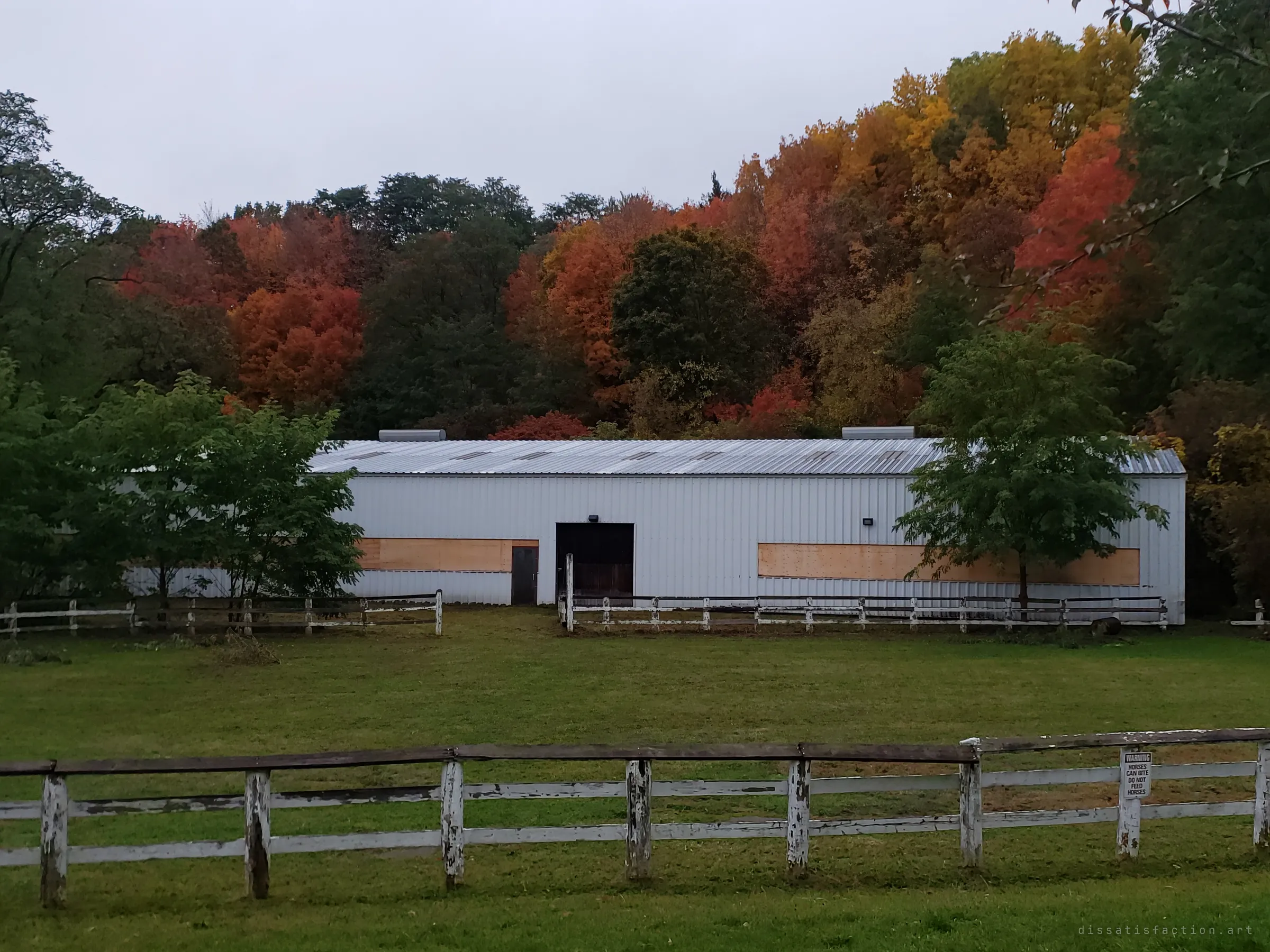 A long white metal barn with boarded windows sits behind a peeling paddock fence, backed by a hillside of red and orange autumn trees. The first look at the stables, shut up and quiet.