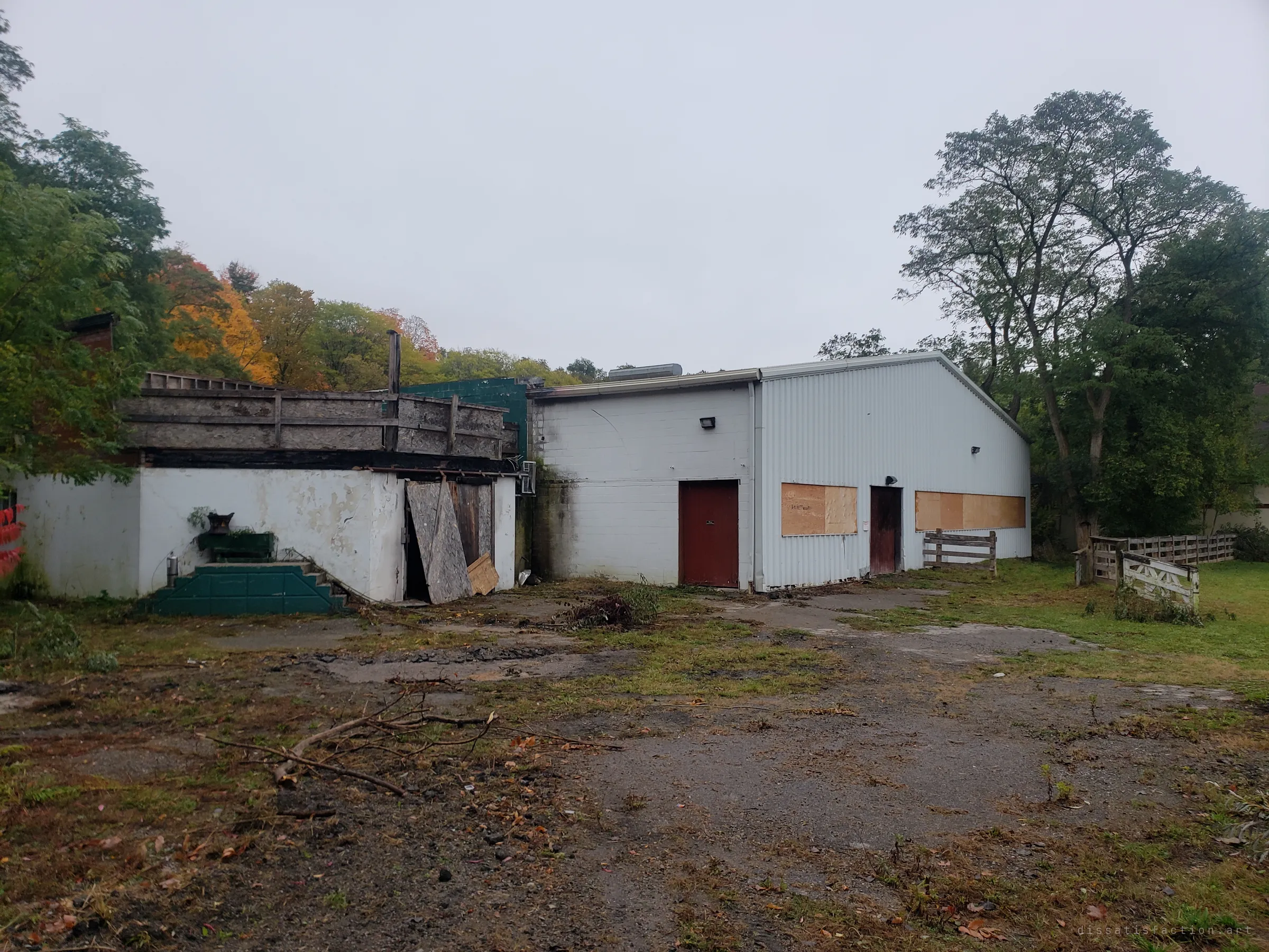 A weedy, cracked yard leads to a white block annex with a collapsing plywood door and a boarded-up metal barn beyond. Branches and debris lie where horses and trailers once came and went.