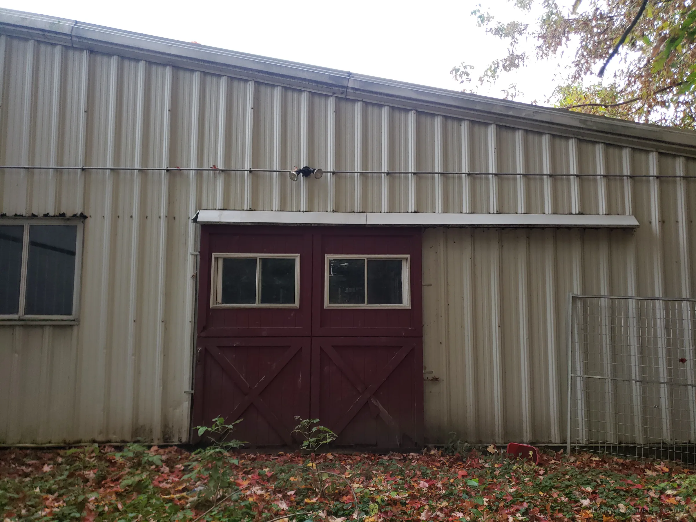 Red cross-braced barn doors with small windows set into a stained metal wall, fallen leaves and ivy creeping up to the sill. A side entrance nobody has used in a while.