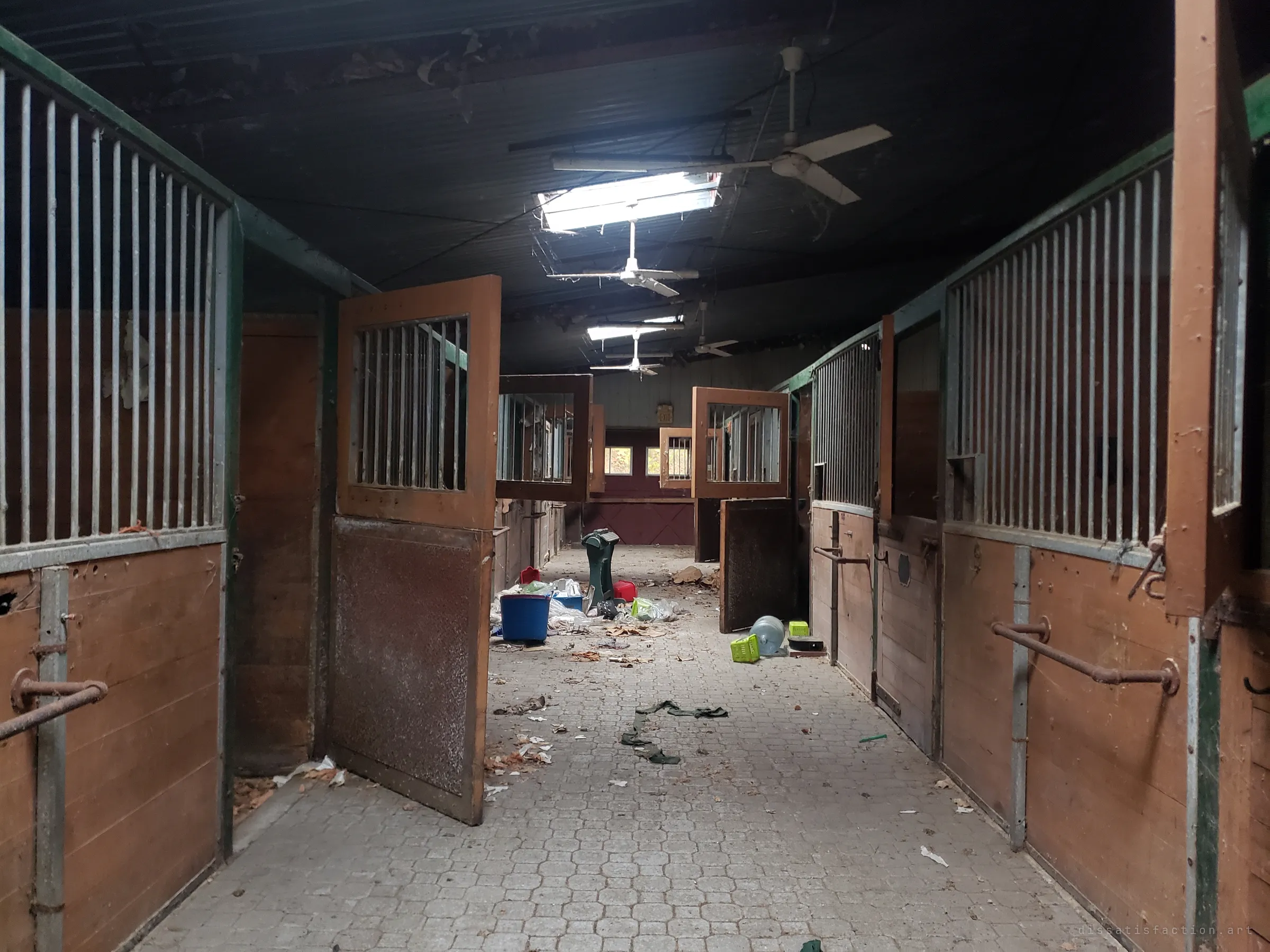 The stable aisle stretches between rows of open box stalls with barred upper doors, ceiling fans still hanging under a broken skylight. Buckets, feed bins and litter are scattered across the paver floor.