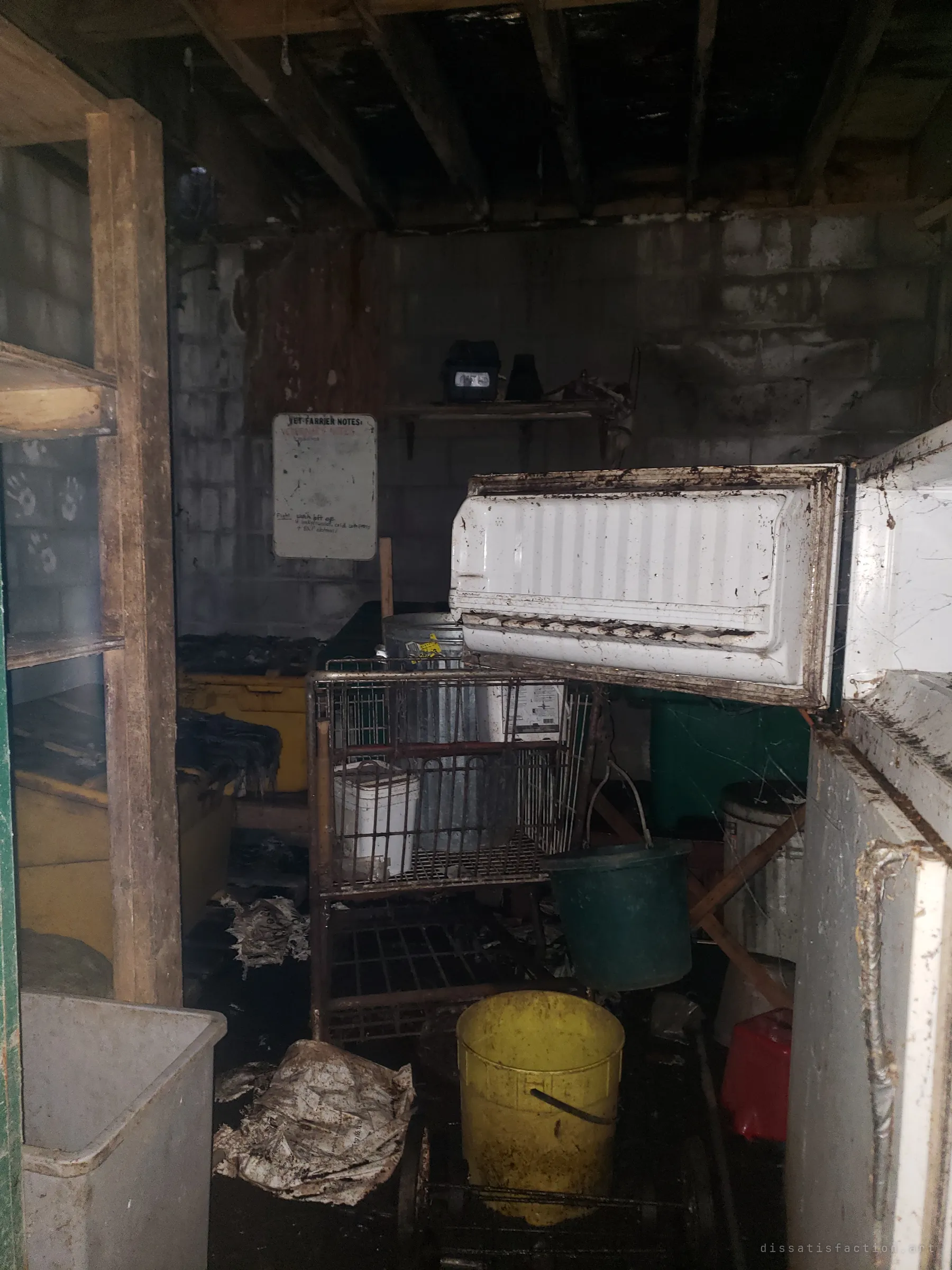 A dark, grimy storeroom packed with an open fridge, a rusted cart, buckets and bins, and a faded notes whiteboard on the block wall. Everything is coated in dirt and cobwebs.