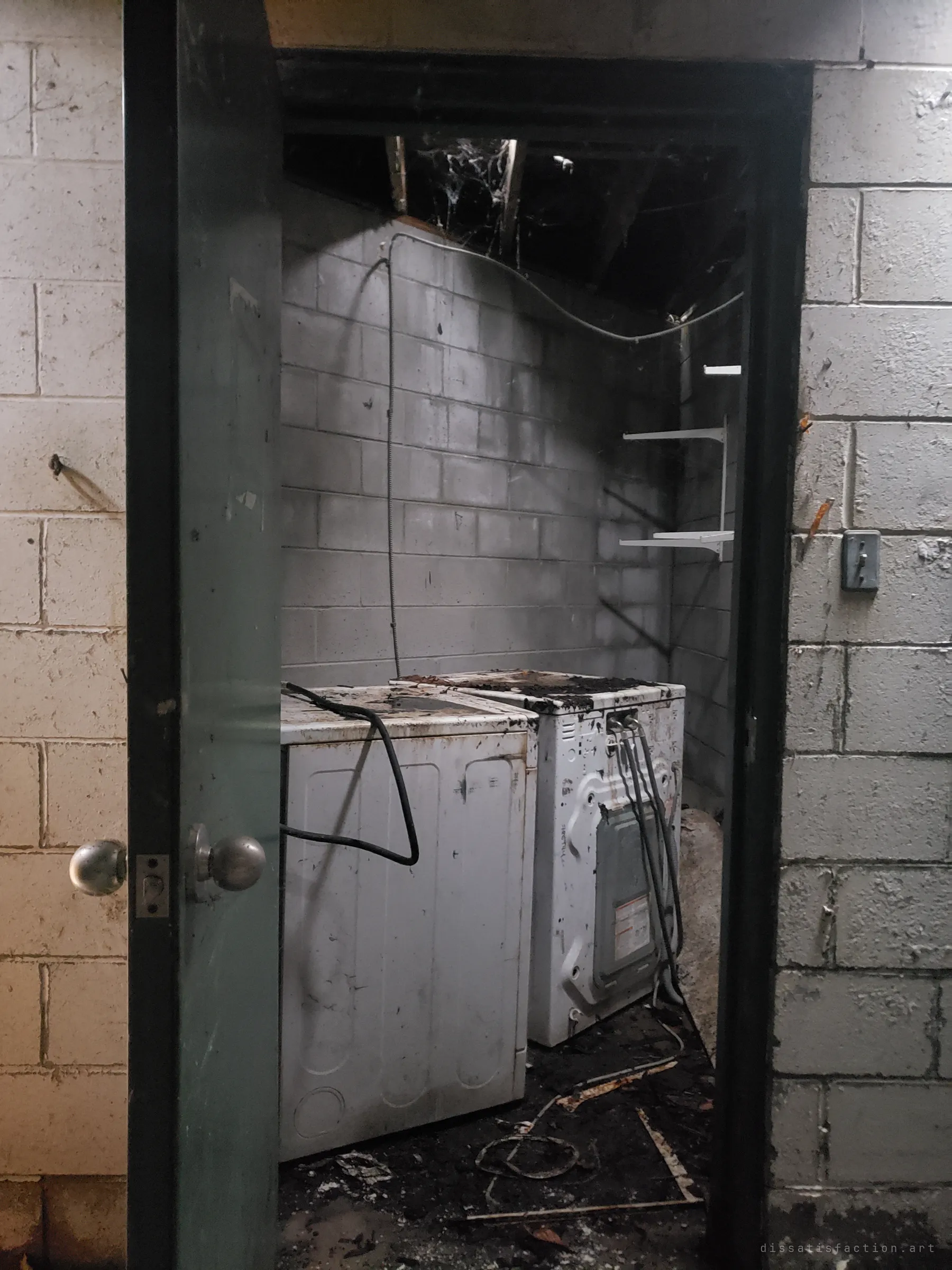 Through a green door, a soot-stained laundry room holds two filthy washing machines, loose cables and bare shelves. The ceiling above is opened up and hung with cobwebs.