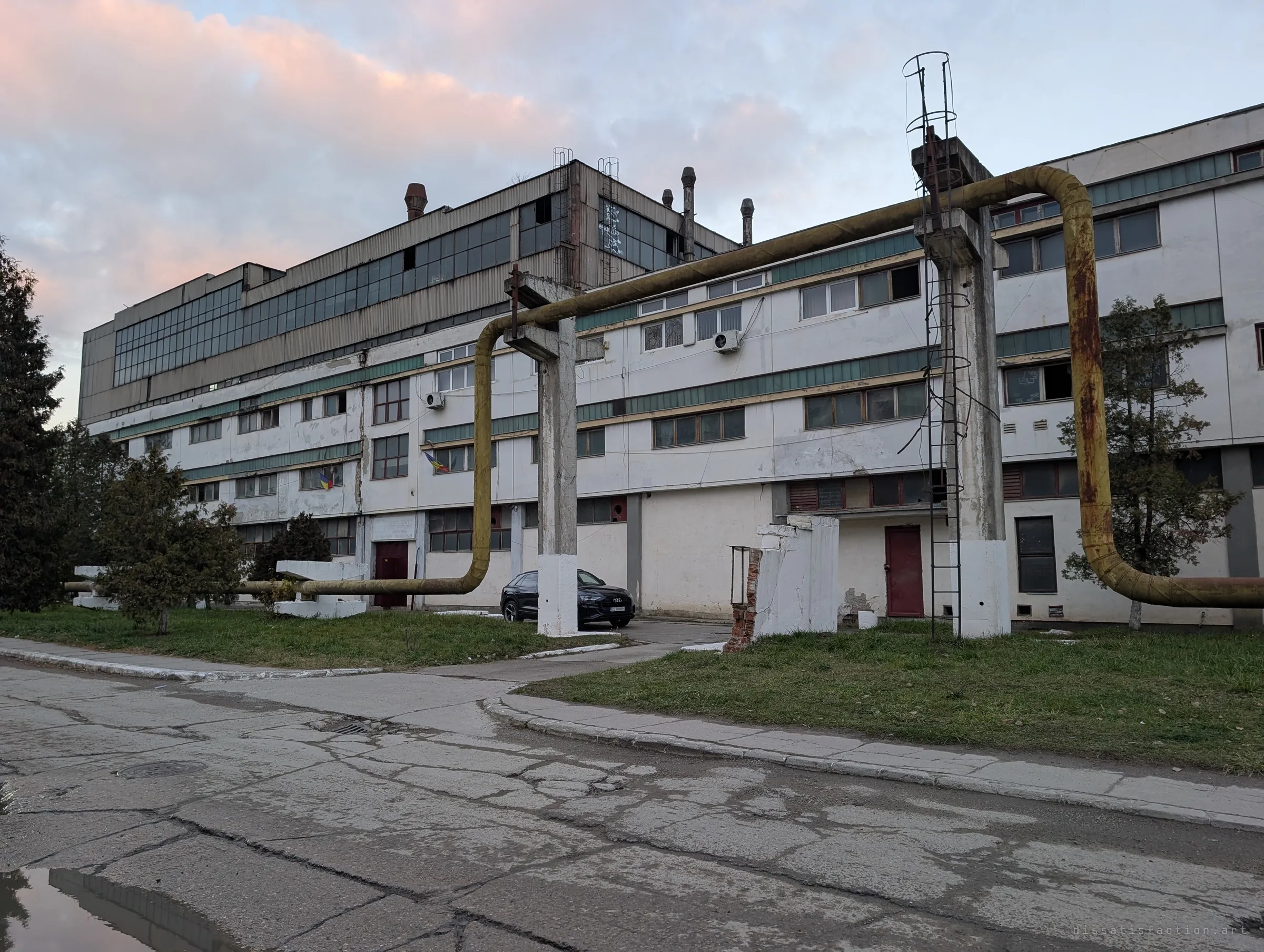 A white multi-storey factory block at dusk, crossed by a rusty yellow overhead pipe on concrete supports, with a parked car and national flags showing part of it is still in use.