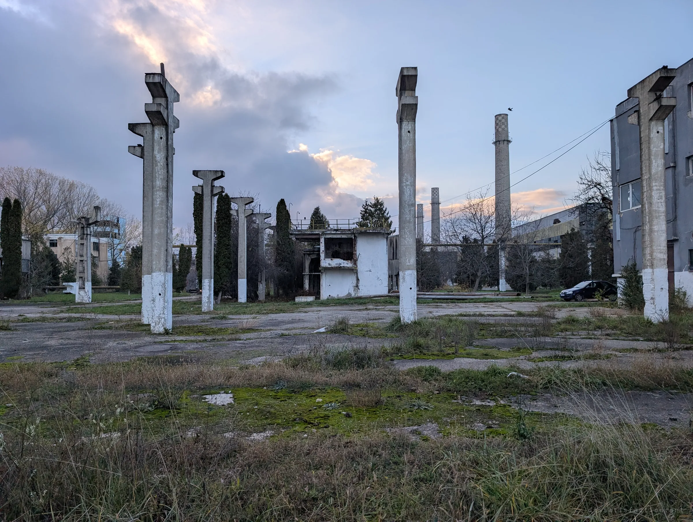 Bare concrete columns stand in a weedy, cracked yard where a hall once stood, with tall chimneys and a gutted control building beyond under an evening sky.
