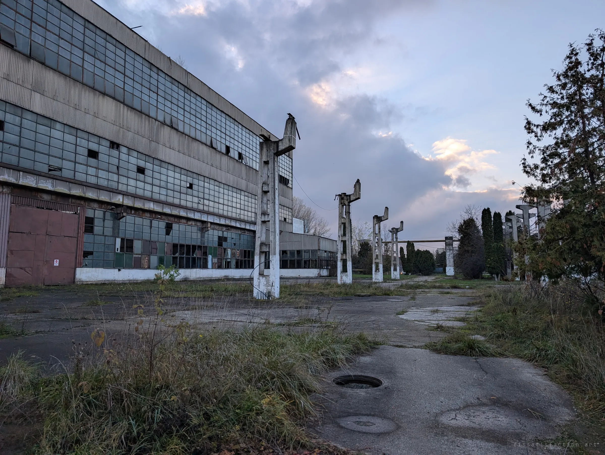 The long glazed wall of a derelict hall runs off to the left while a line of broken concrete crane posts leads across the overgrown yard at dusk.