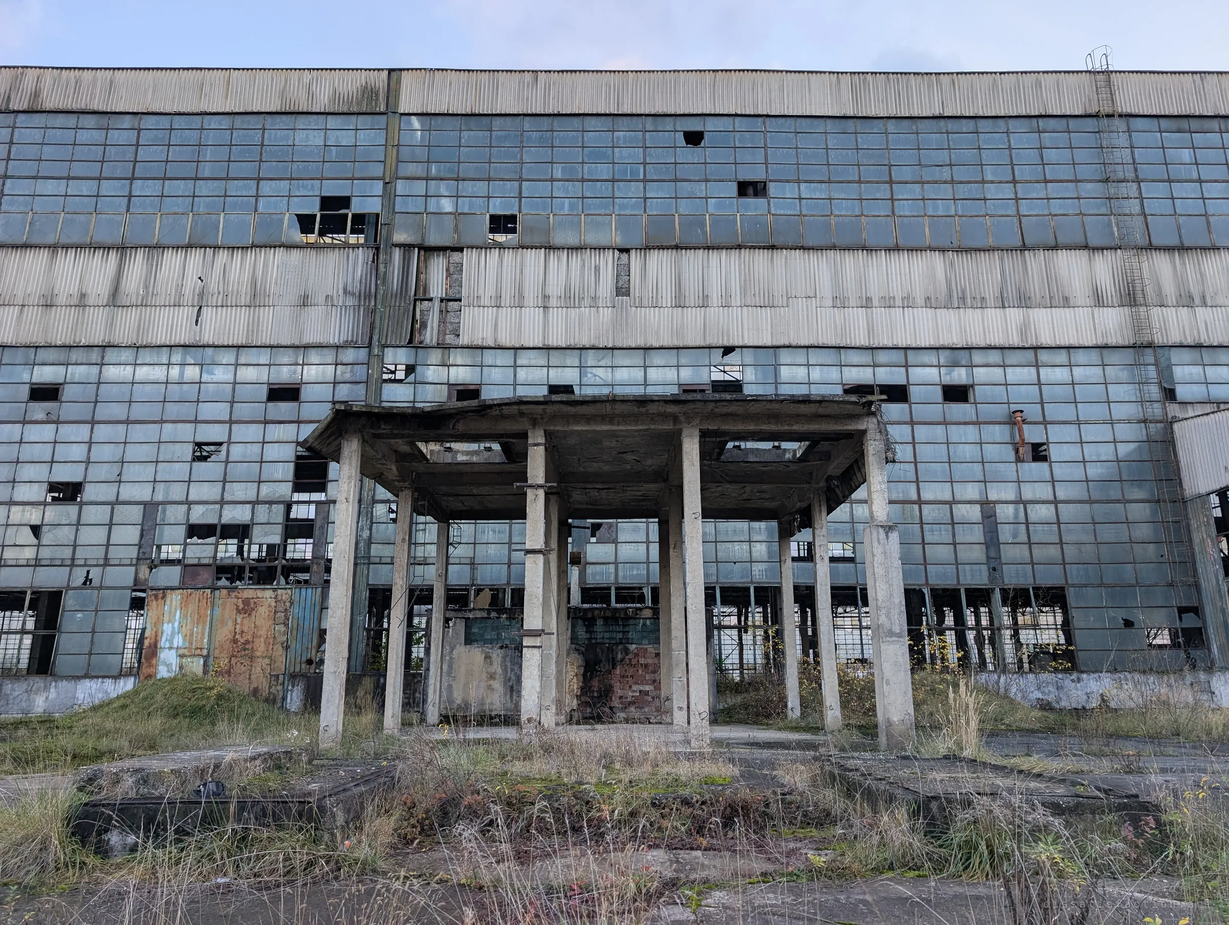 Head-on view of a vast facade of broken glass panes and corrugated cladding, fronted by a crumbling concrete canopy on thin pillars.
