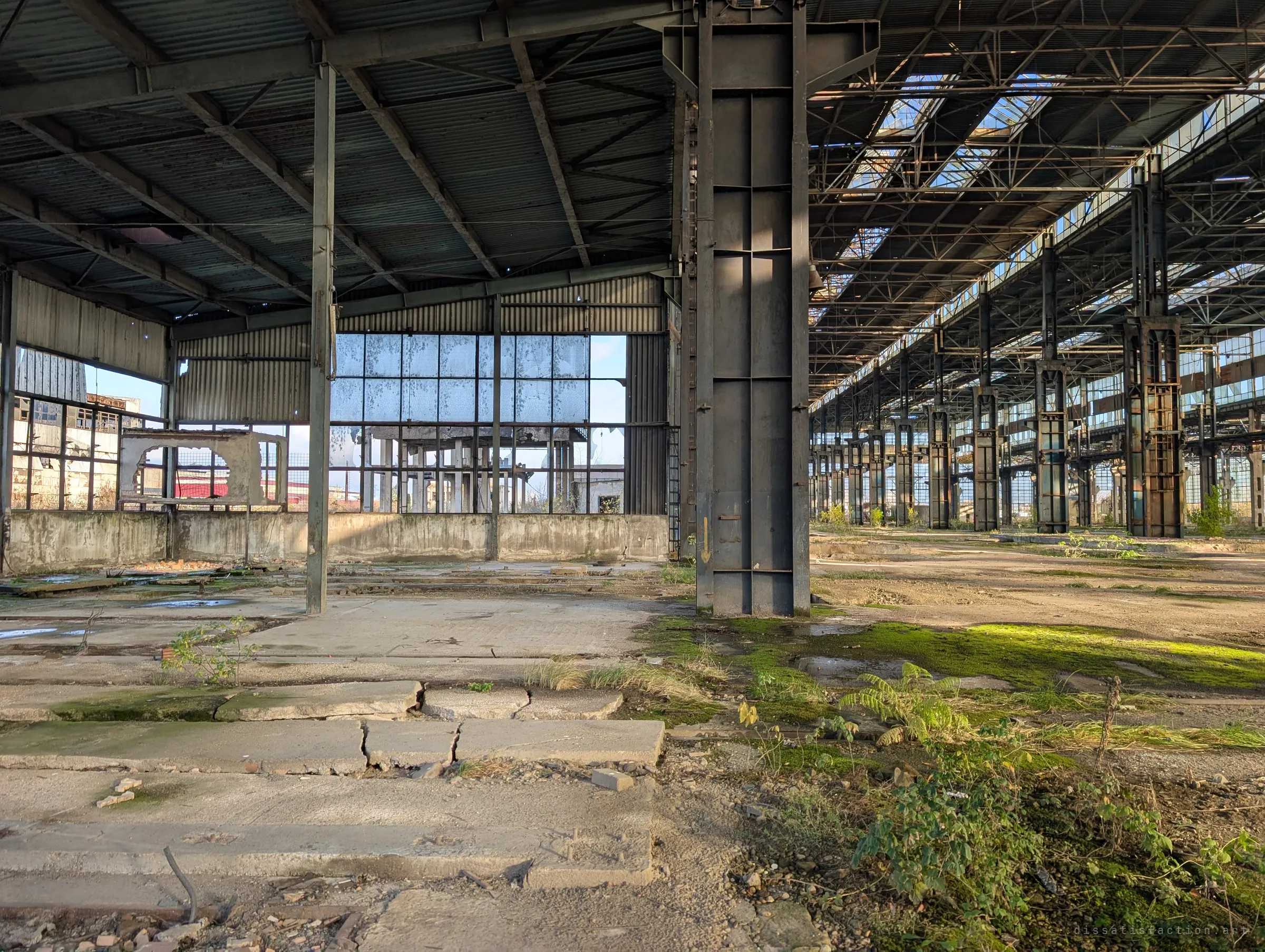 A giant steel column splits the view between a side bay with a torn-open glass wall and the long main hall receding into the distance.
