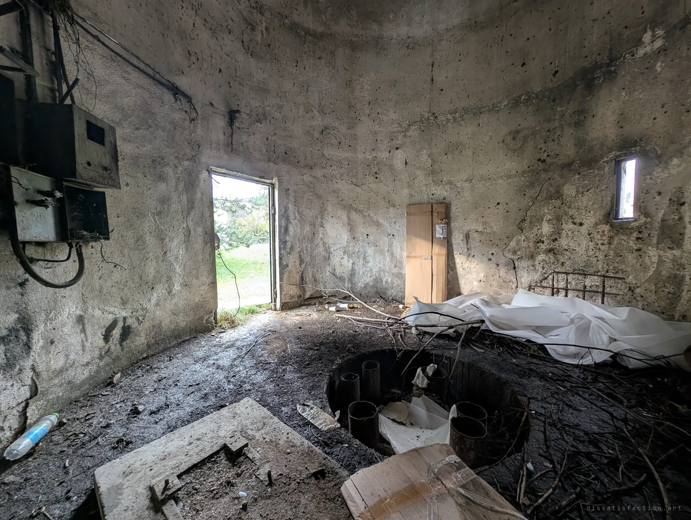 The round concrete base room of the tower, with dead electrical boxes on the wall, an open door onto greenery and a pit of pipe stubs choked with branches and plastic sheeting.