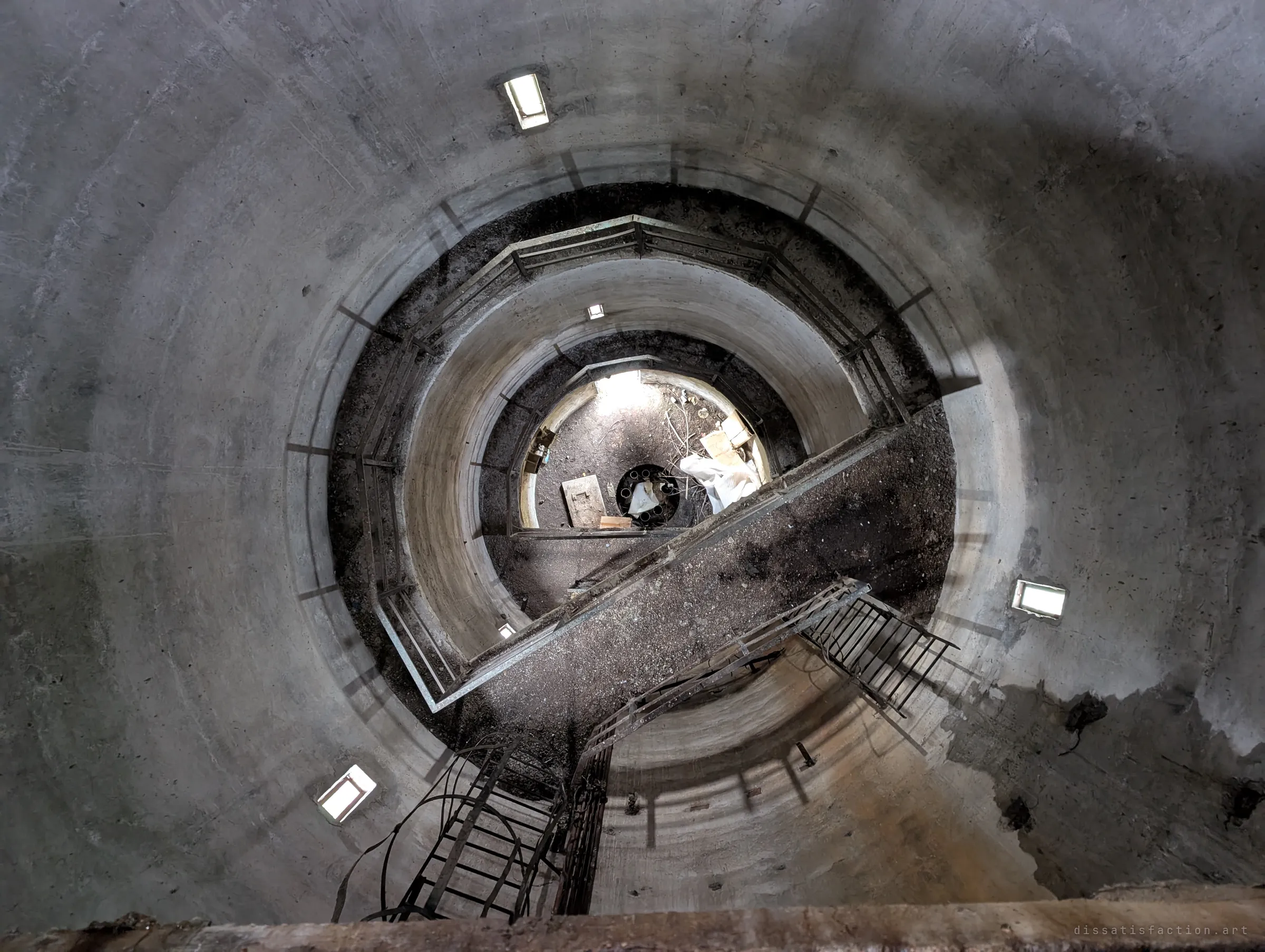 Looking straight down the tower's hollow shaft through concentric concrete rings, railings and a steep stair to the littered floor far below. The dizzying depth is the point.