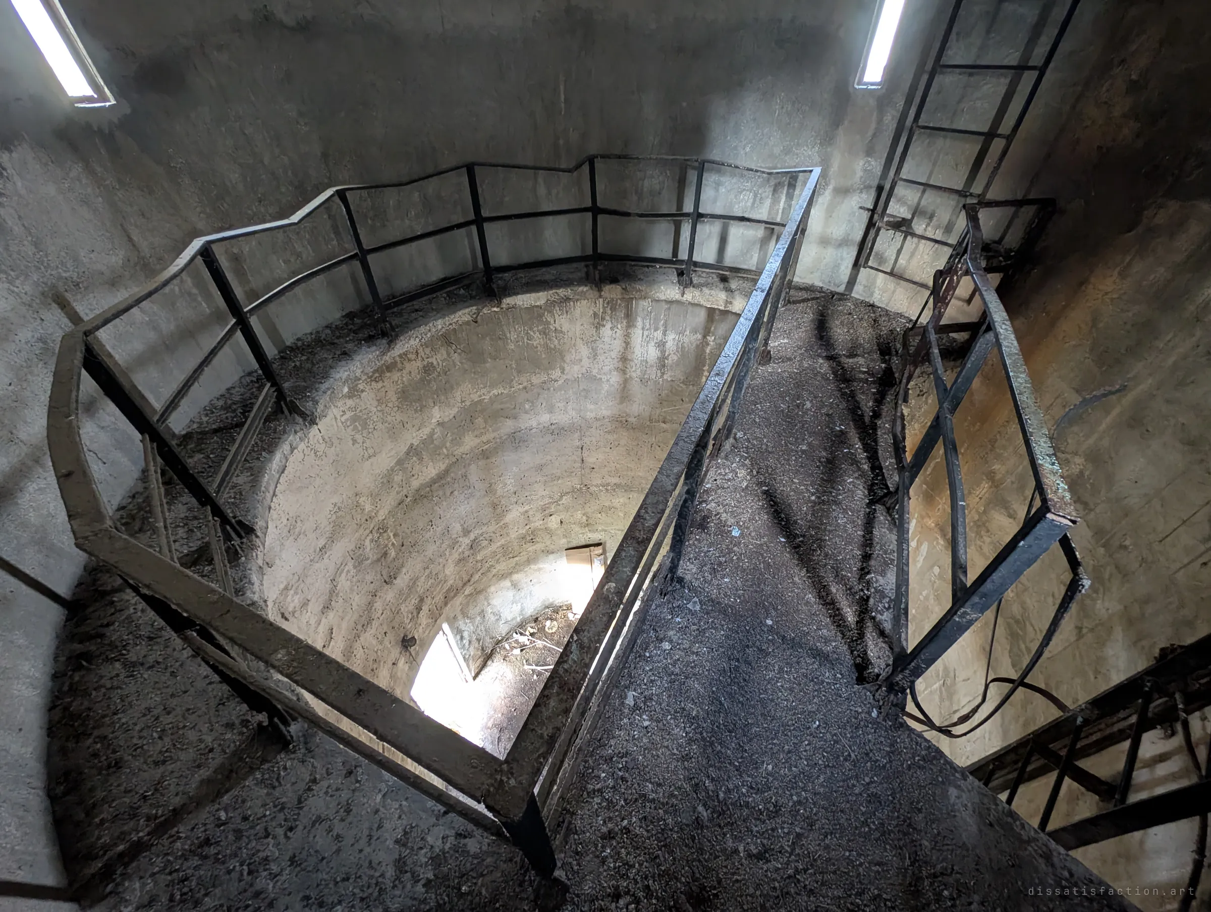 A railed landing inside the tower shaft, with a narrow walkway crossing the drop and a ladder climbing past slit windows. Grit and debris cover every surface.