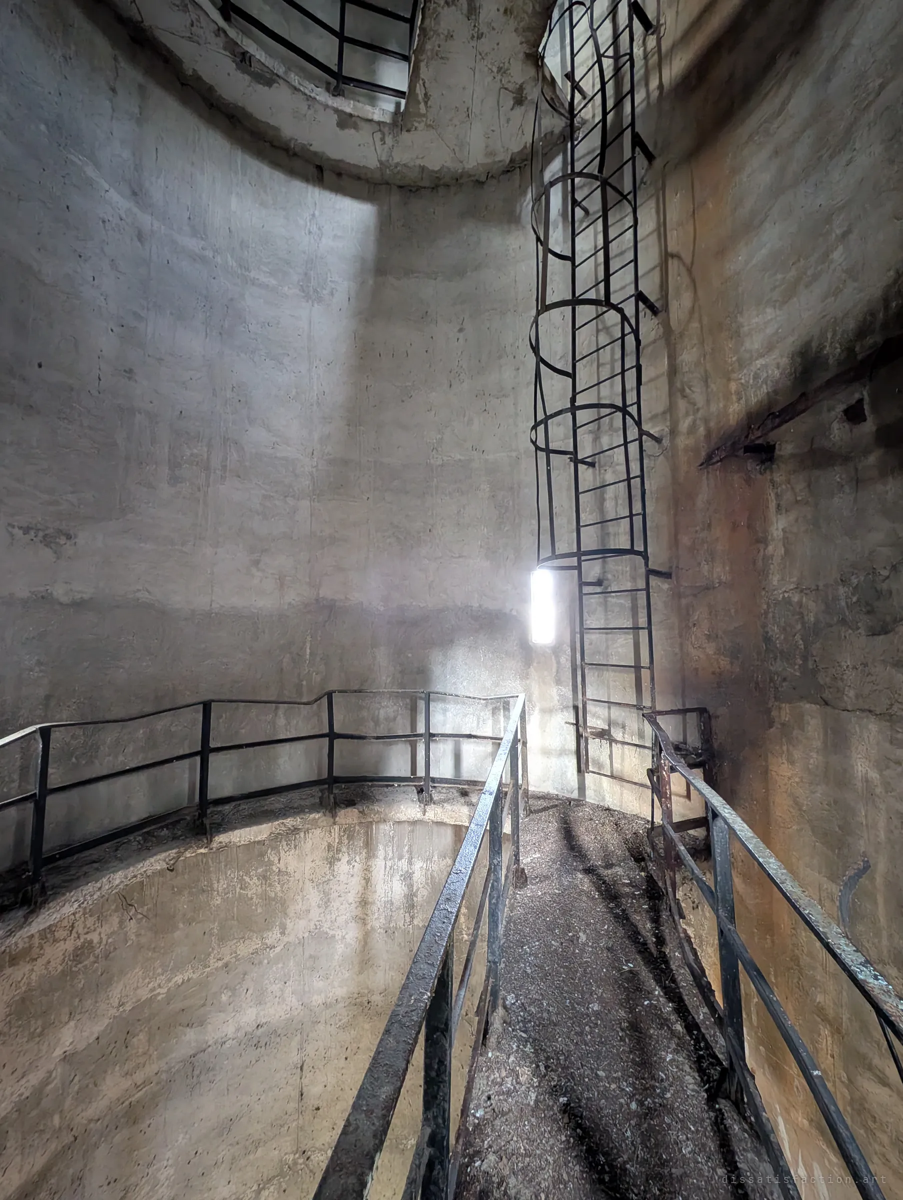 Inside the round concrete shaft of the water tower, a narrow railed walkway crosses above a drop while a caged ladder climbs toward the light. The single slit window throws a hard beam across the curved wall.