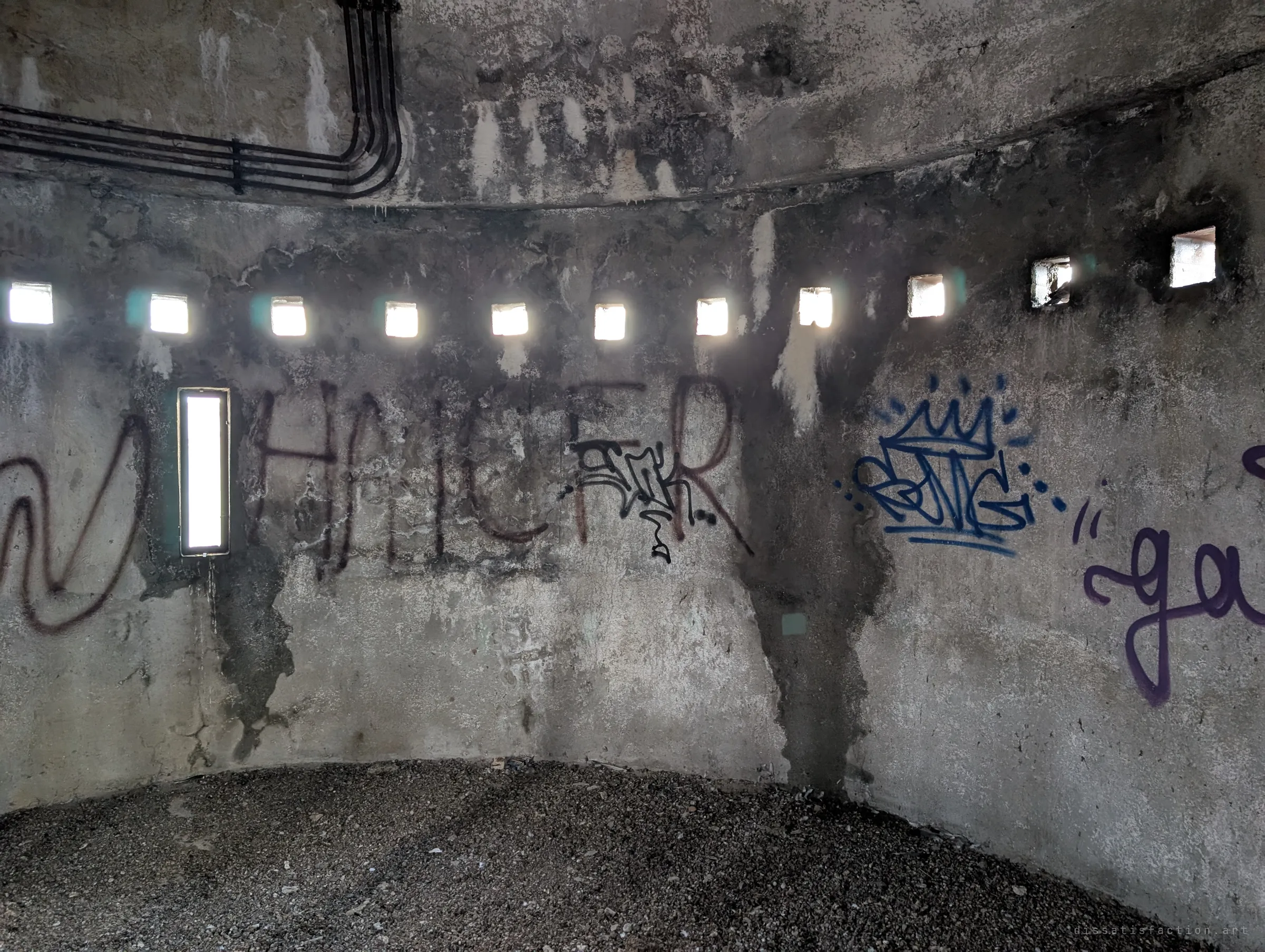 A circular room near the top of the tower, ringed by small square windows and one tall slit, its walls covered in layered graffiti tags. Gravel and debris cover the floor.