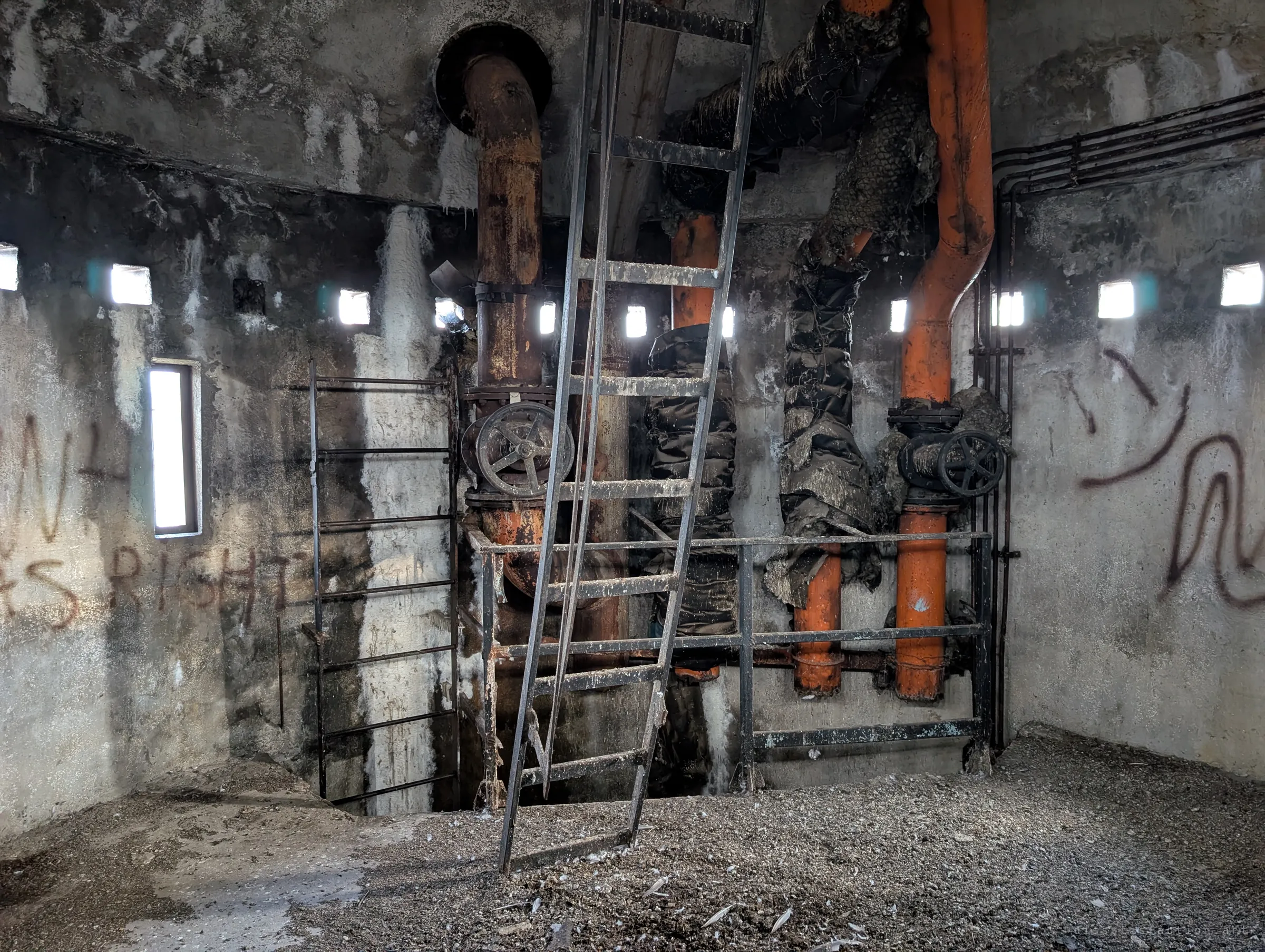The tower chamber around the main risers: rust and orange pipes with handwheel valves, torn insulation and a loose ladder leaning over the open shaft.