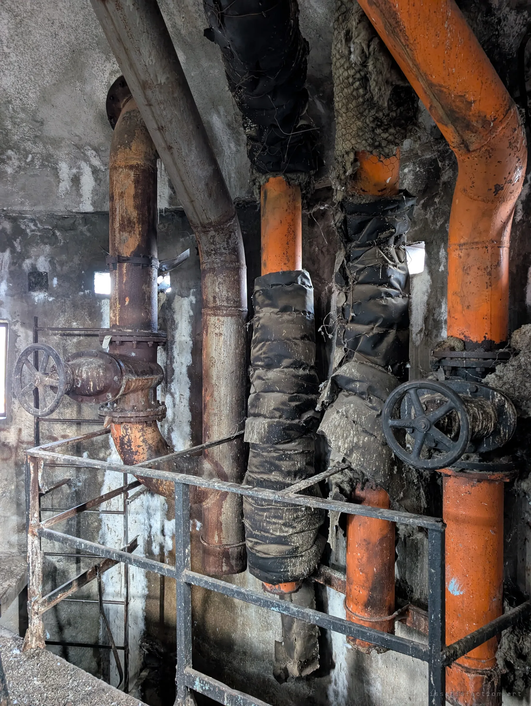 Close view of the tower risers, orange-painted and rusted pipes wrapped in shredded black insulation, with two large handwheel valves behind a railing.