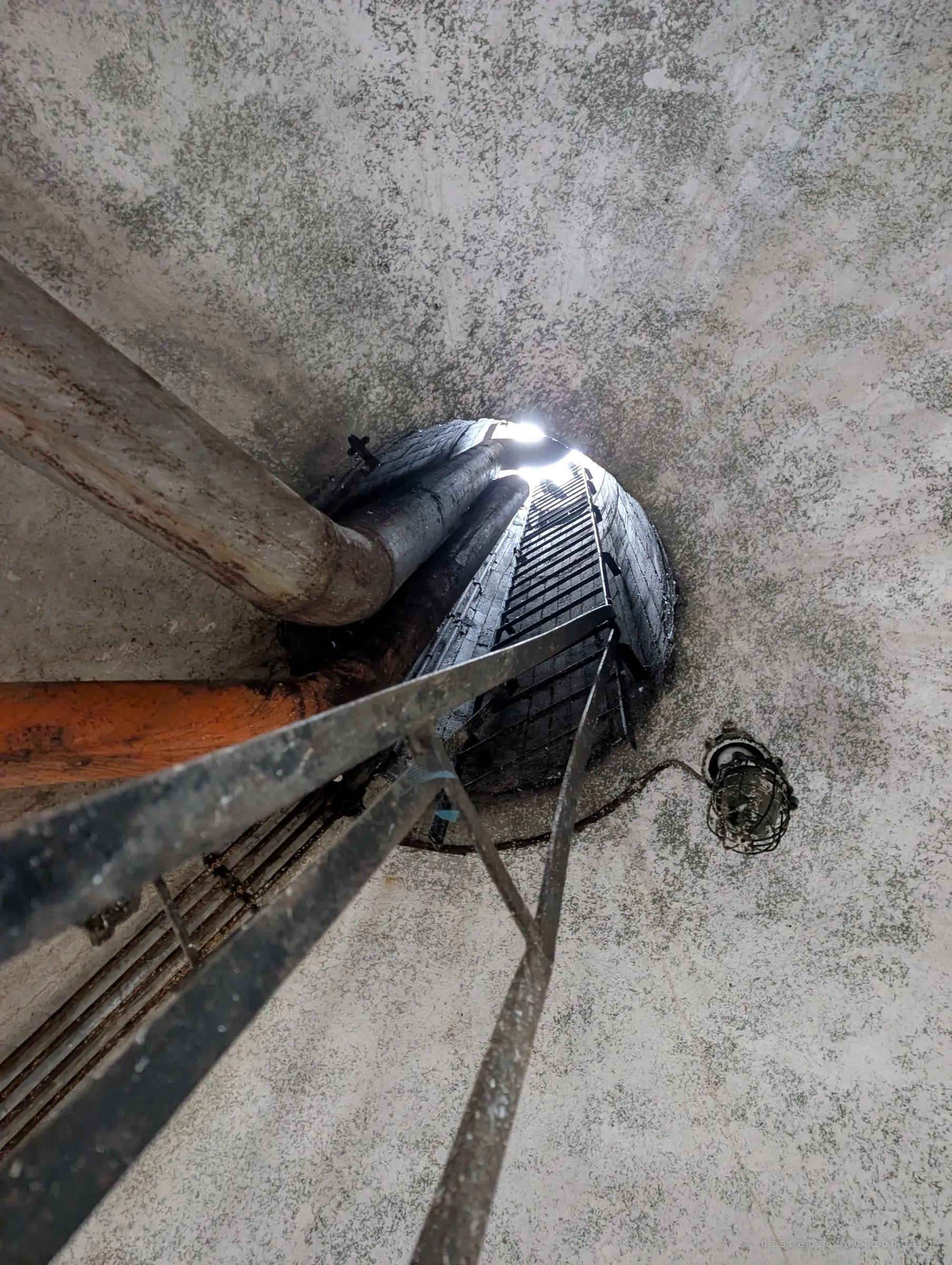 Looking straight up a narrow brick-lined shaft, a long ladder rising past pipes toward a bright opening. An old caged lamp hangs on the concrete ceiling beside it.