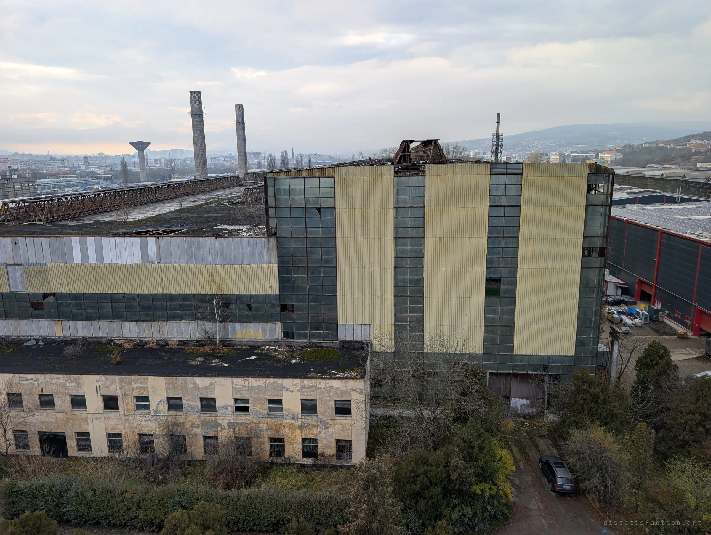 From the top of the tower, the tall glazed hall and its low office block stretch out below, with chimneys and a distant city under a grey sky. It shows the scale of the complex in one frame.