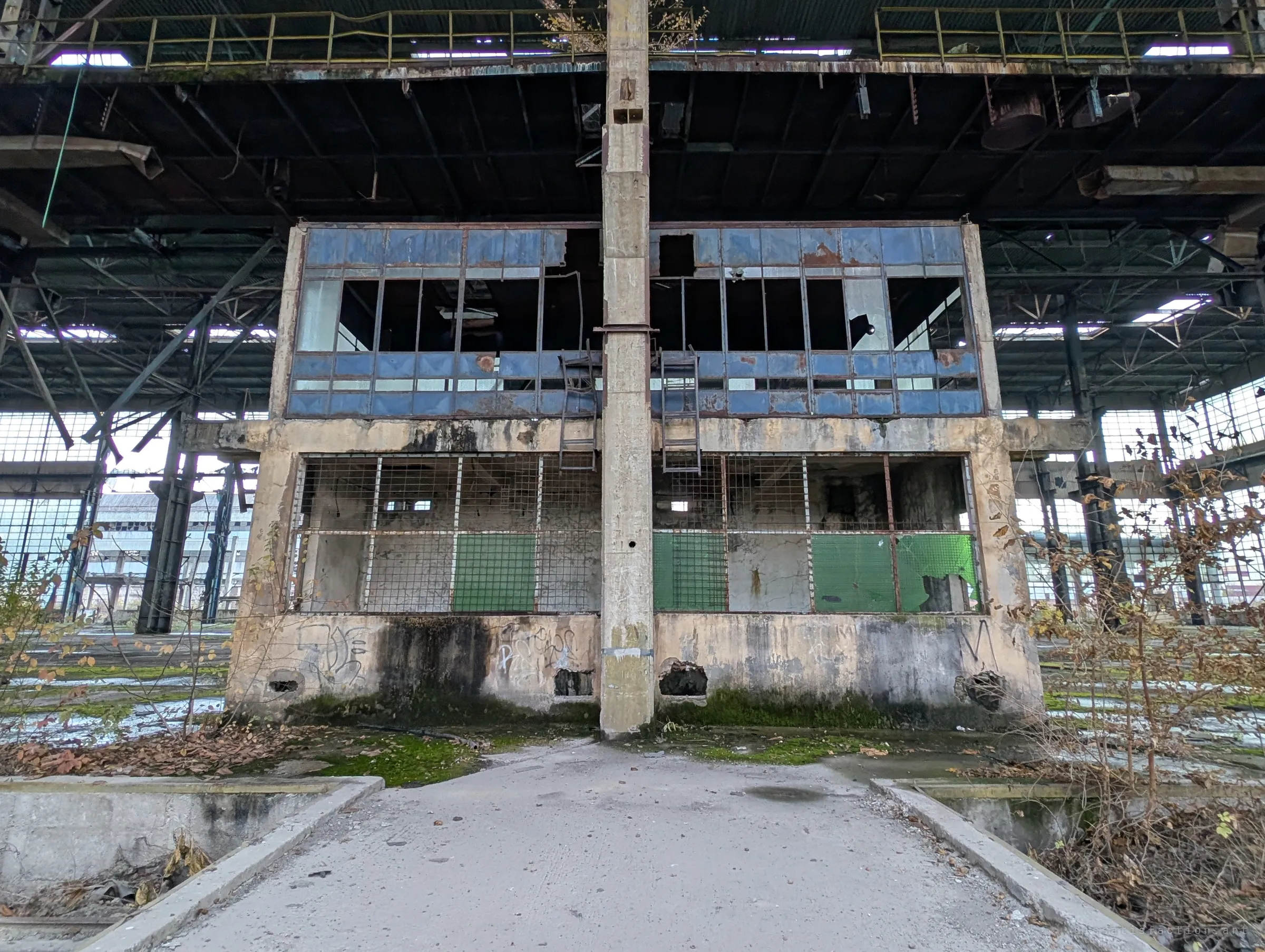 A two-storey control booth with shattered blue glazing stands in the middle of a vast hall, a concrete column splitting it down the centre. Moss creeps across the floor where heavy machinery once moved.