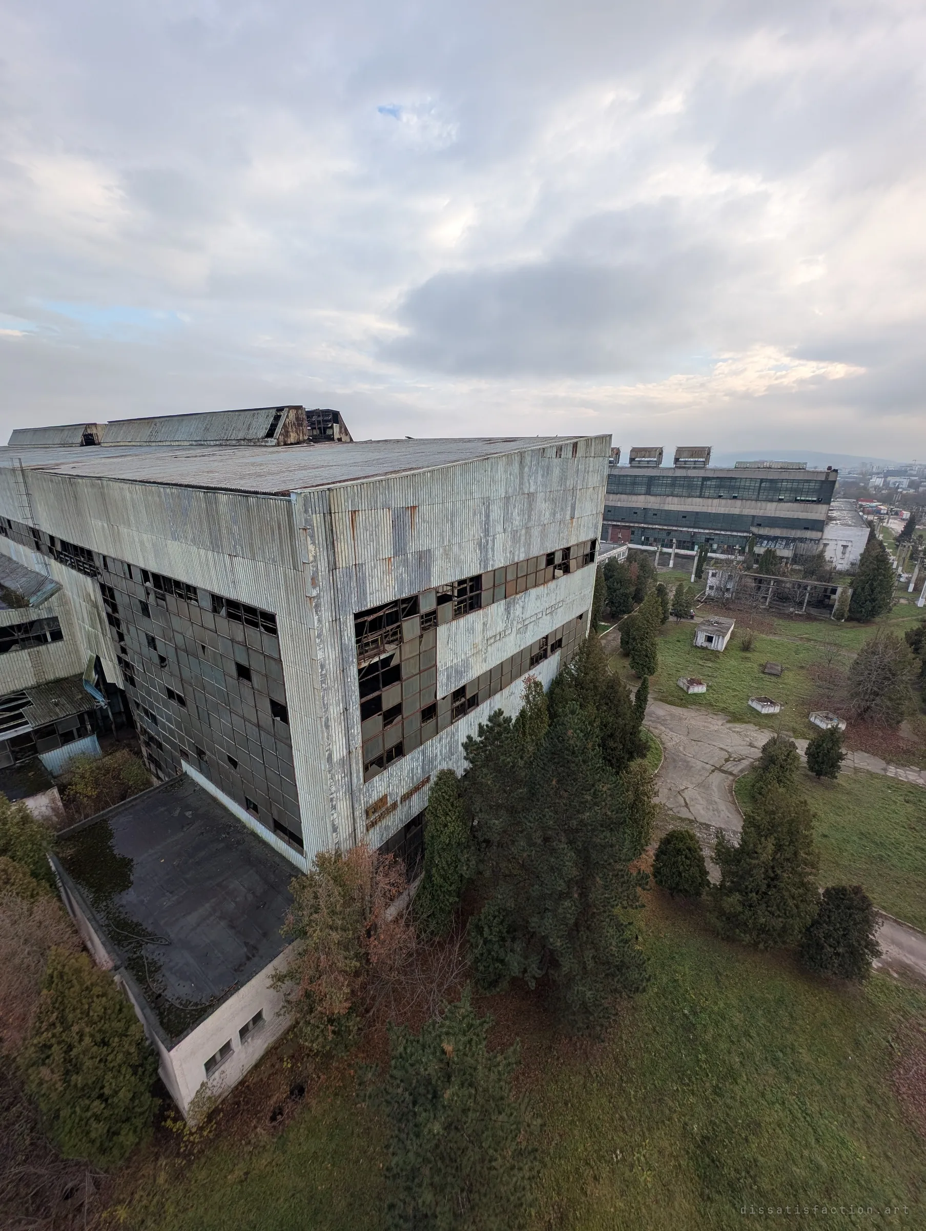 Looking down from the tower onto a huge corrugated hall with shattered window bands, pines crowding its base and more halls beyond. Faded department lettering still clings to the facade.