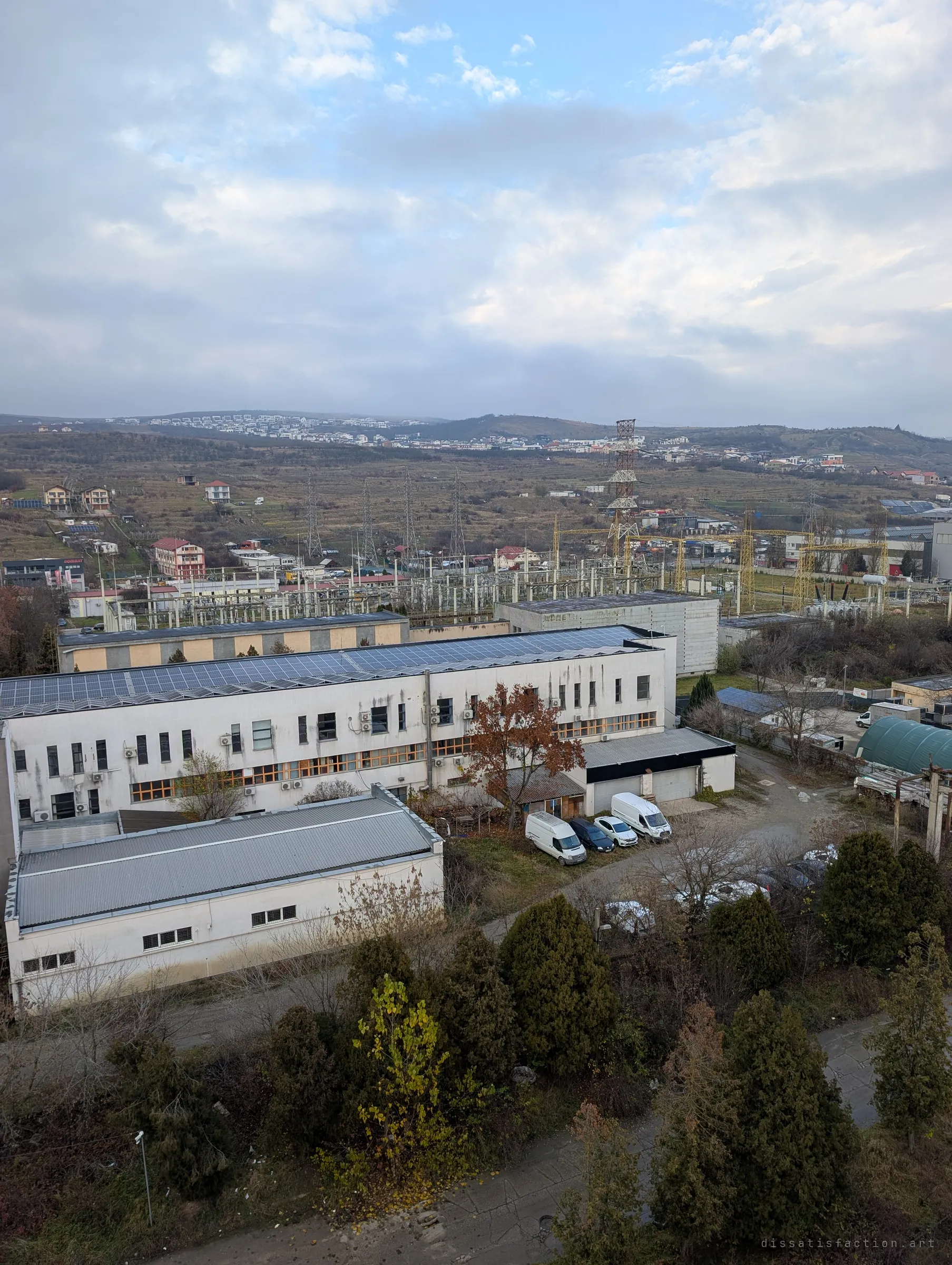 The view out from the tower over an active neighbouring building with rooftop solar, a substation and pylons, and houses spreading up the hills. It shows how the ruin sits inside a living district.