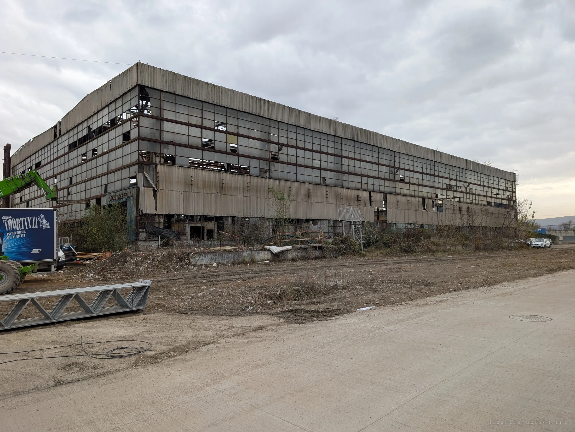 One of the long production halls seen from a cleared yard, its multi-storey window grid mostly smashed and a foundry department sign over the entrance. Machinery and a delivery trailer hint at clearance work.