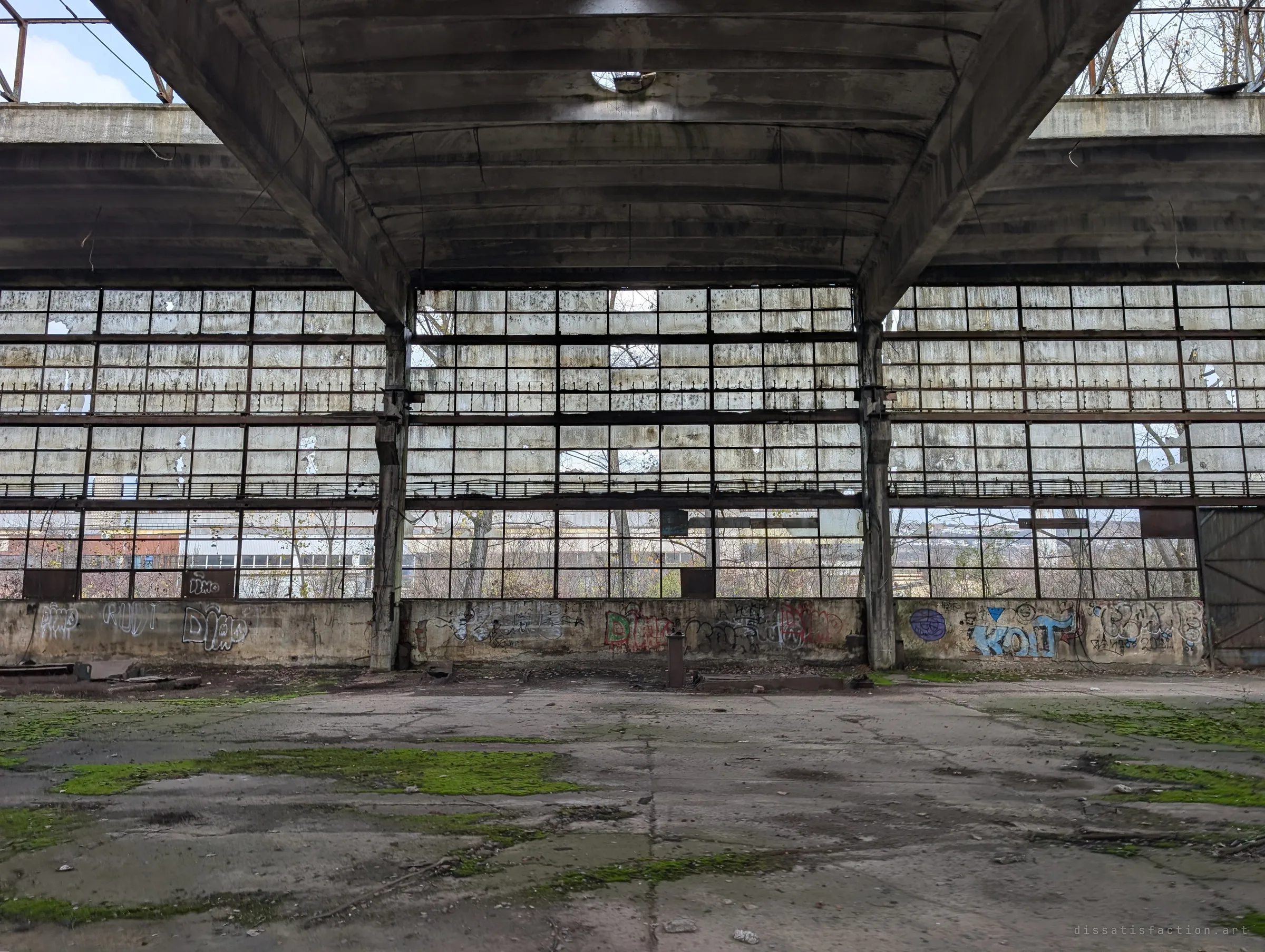 A tall glazed end wall of a hall, most panes cracked or missing, under a heavy concrete roof bay. Graffiti runs along the low wall and moss spreads over the floor.