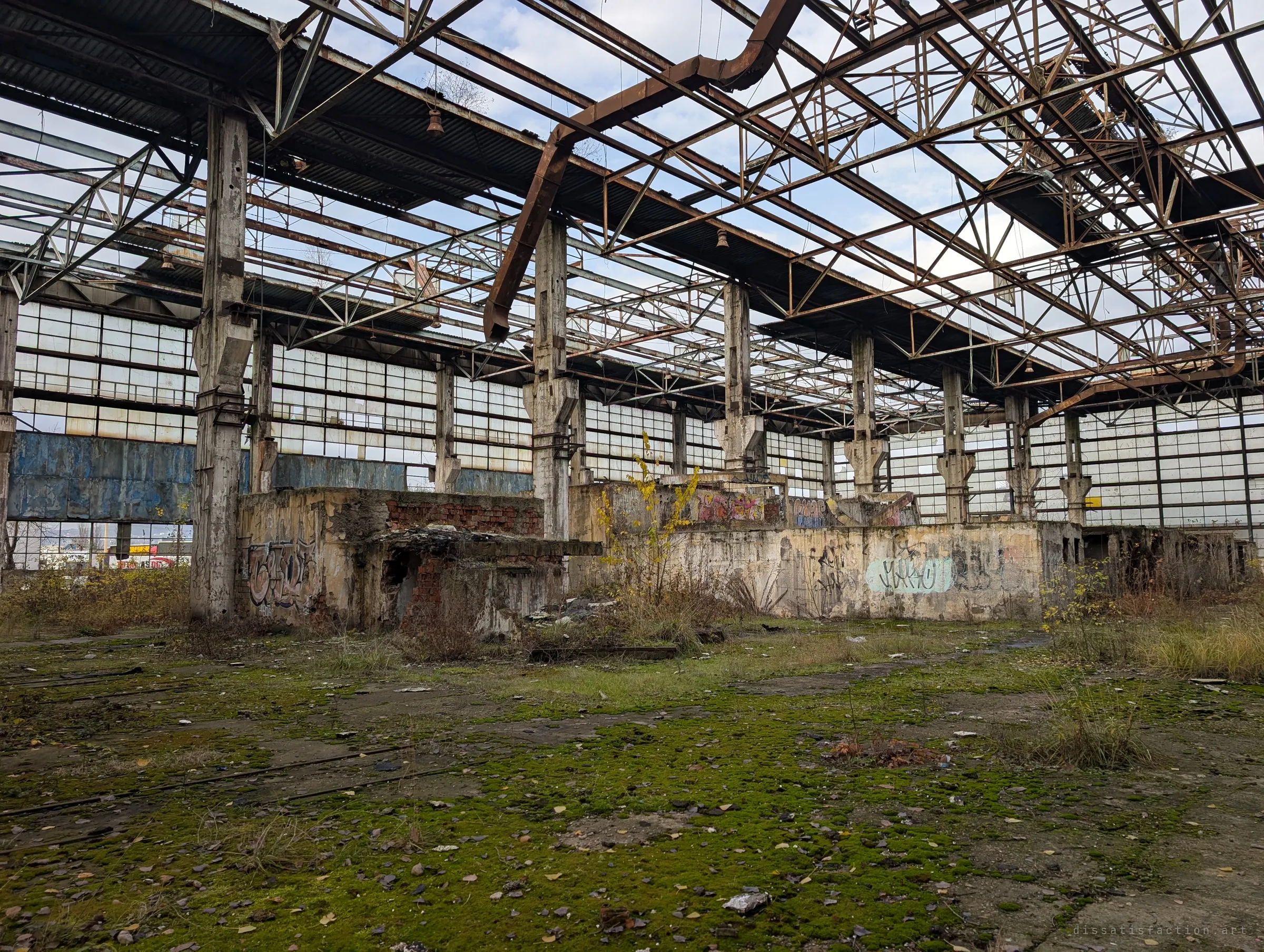 A roofless steel-truss hall with moss spreading across the floor and graffiti on crumbling concrete plinths. The skeletal roof lets daylight pour over the long ribbon windows.