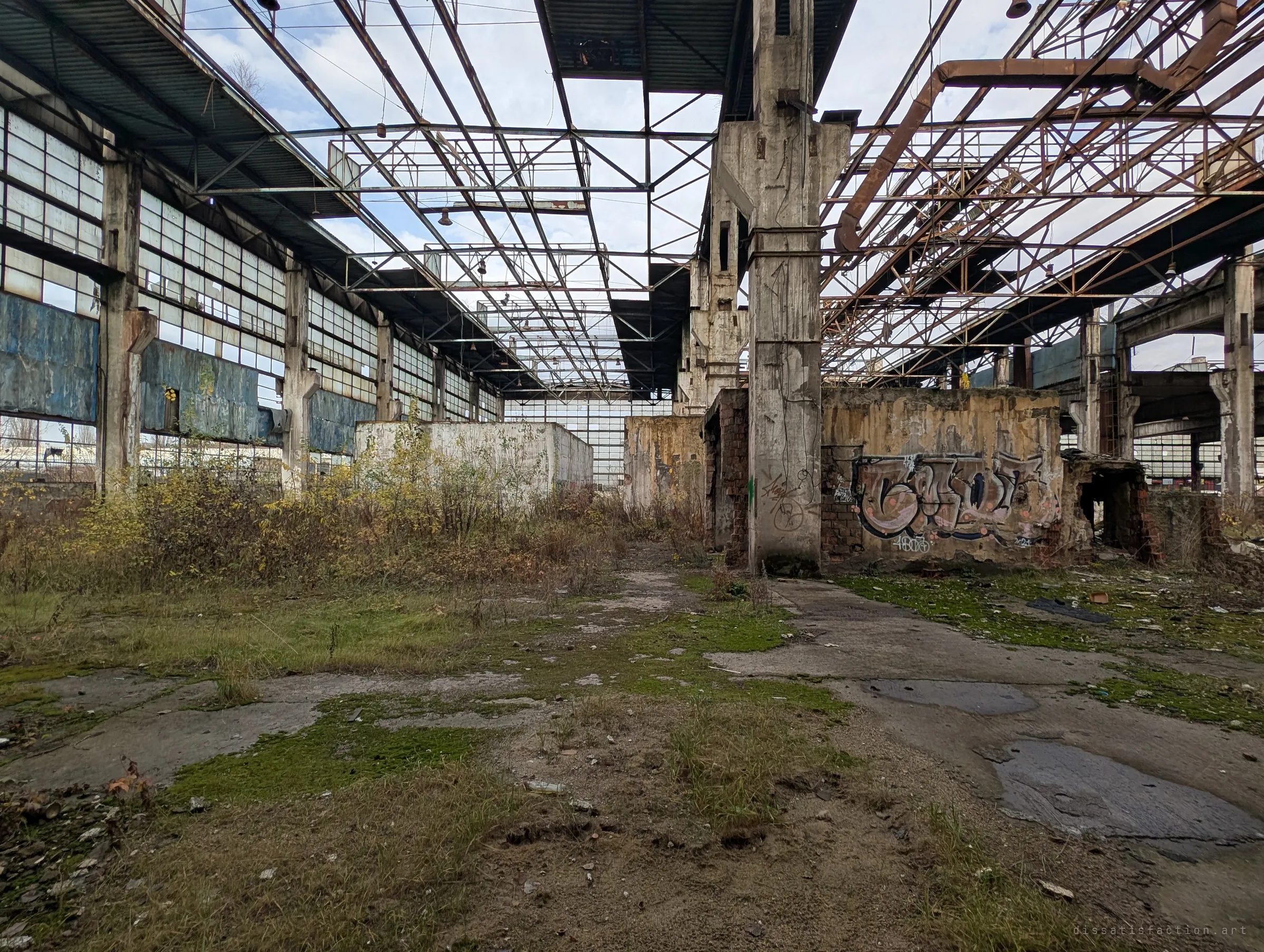 Looking down a weed-choked bay of the hall, open roof trusses above and a big graffiti piece on a brick enclosure. Shows the sheer length of the abandoned production line.