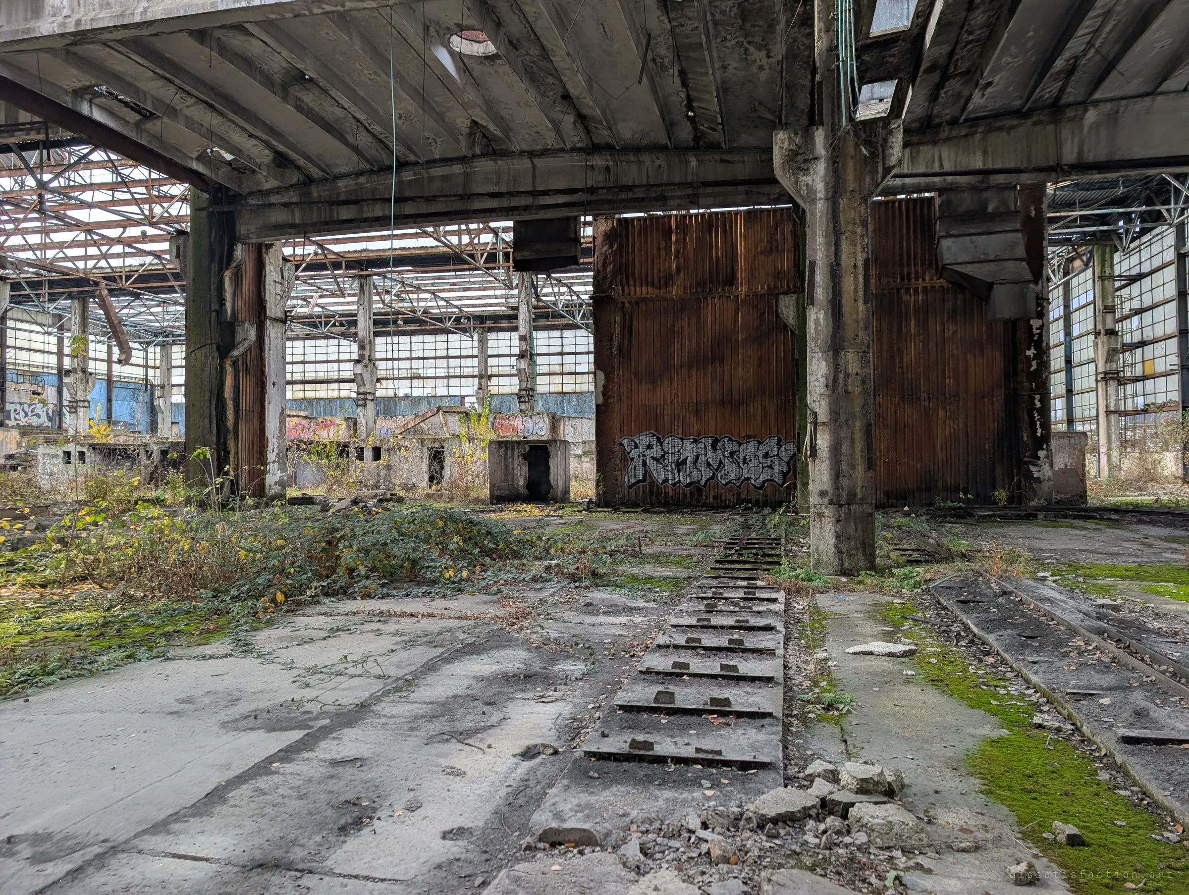 A rusted corrugated wall panel tagged with graffiti stands between concrete pillars, with a slotted rail bed leading toward it. Nature is creeping in over the old workshop floor.