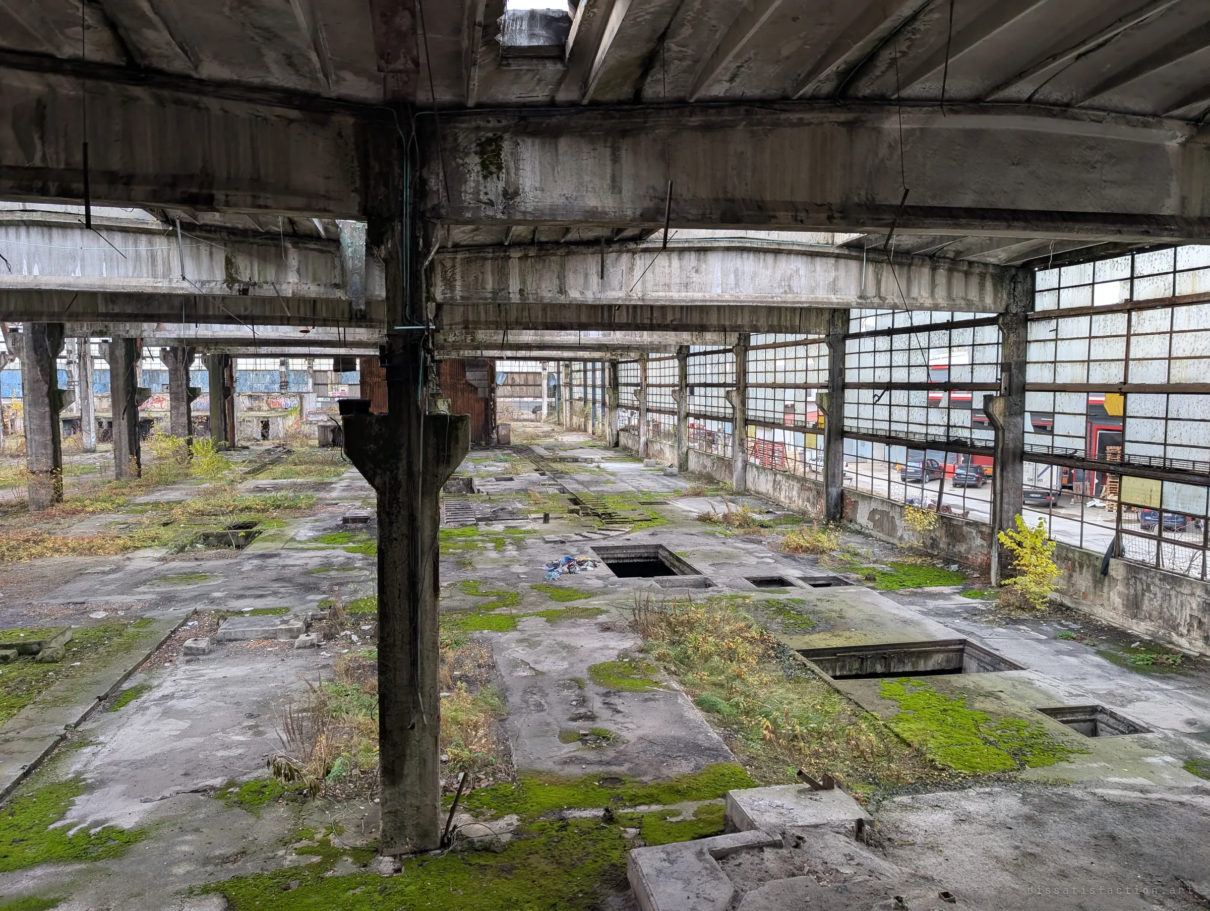 From a raised vantage, a long concrete hall stretches away with open floor pits and moss, while the wall of cracked windows looks out on a still-working yard next door. Dead industry sits right against living business.