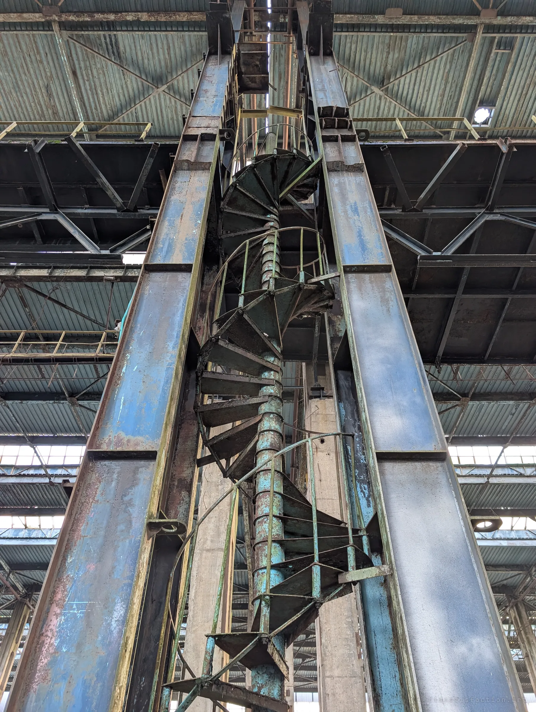 Looking straight up a rusted spiral staircase that winds between two blue steel columns toward a crane gantry under the roof. The peeling paint and scale of the ironwork show how big this plant once was.