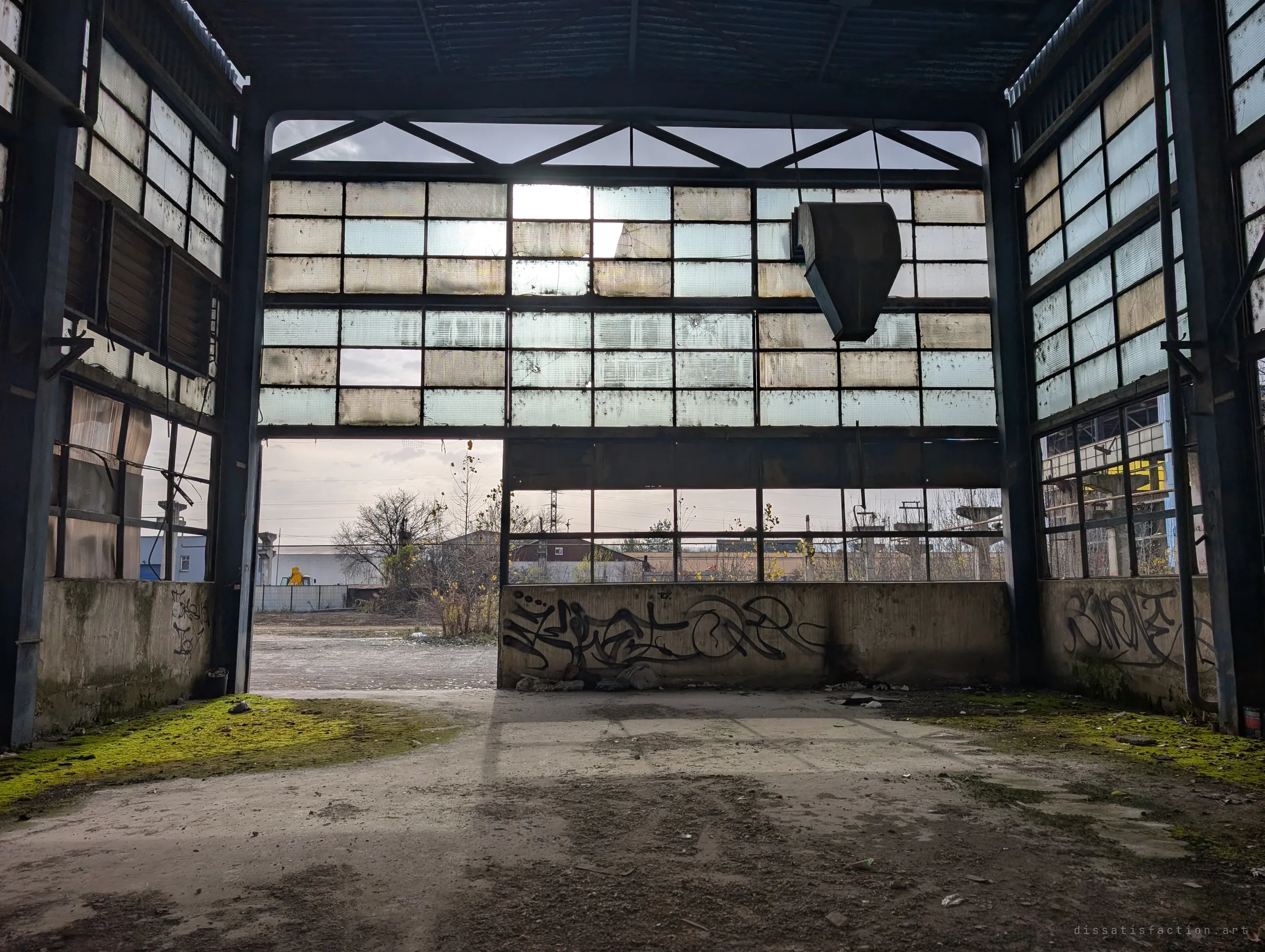 A dark end bay opens onto the grey afternoon through a grid of cloudy glass, a hanging duct silhouetted against the light. The empty yard outside is visible through the gap.