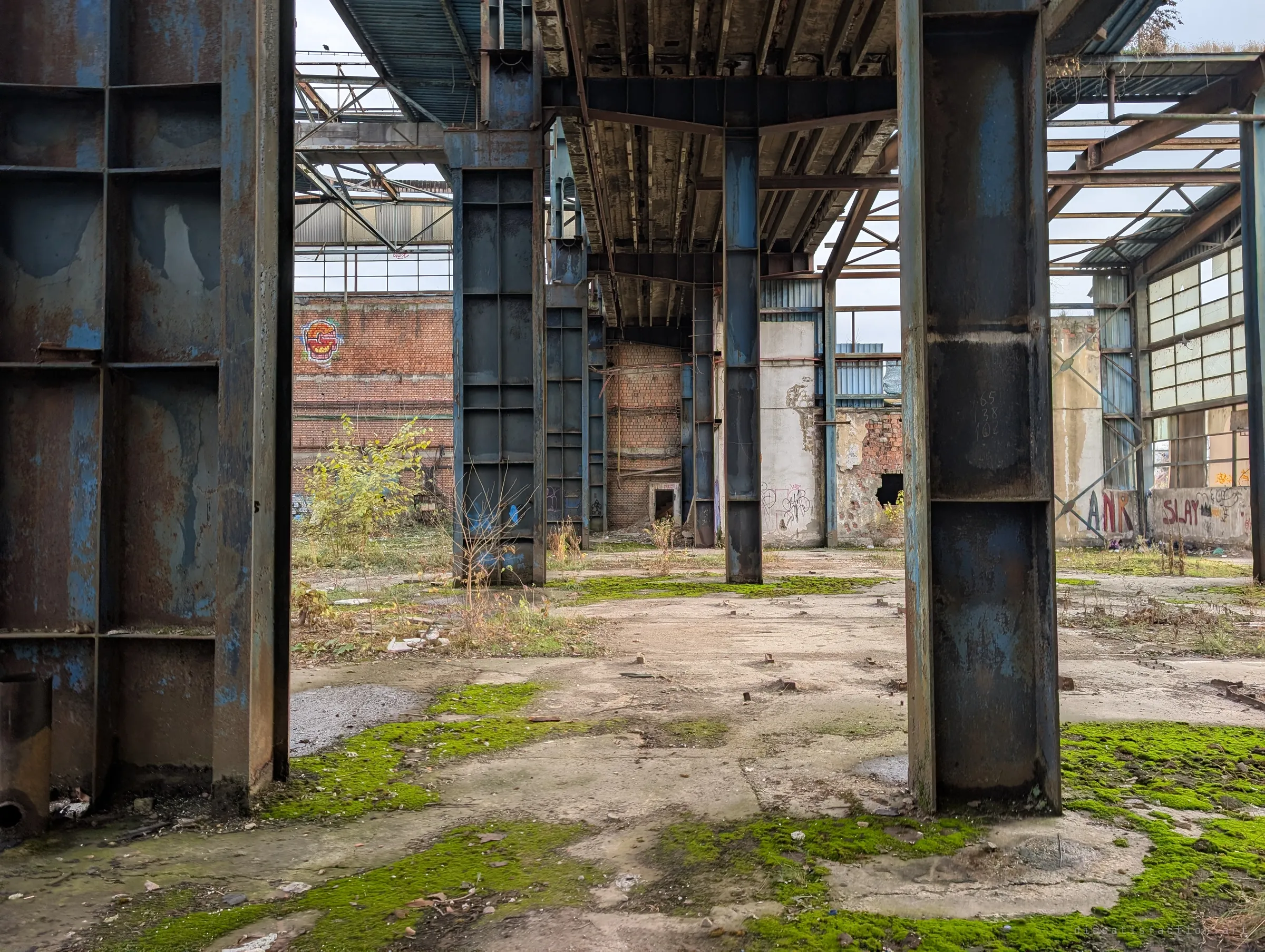 Massive riveted blue steel columns frame a view into further bays of brick and corrugated walls. Bright moss patches break up the concrete floor.