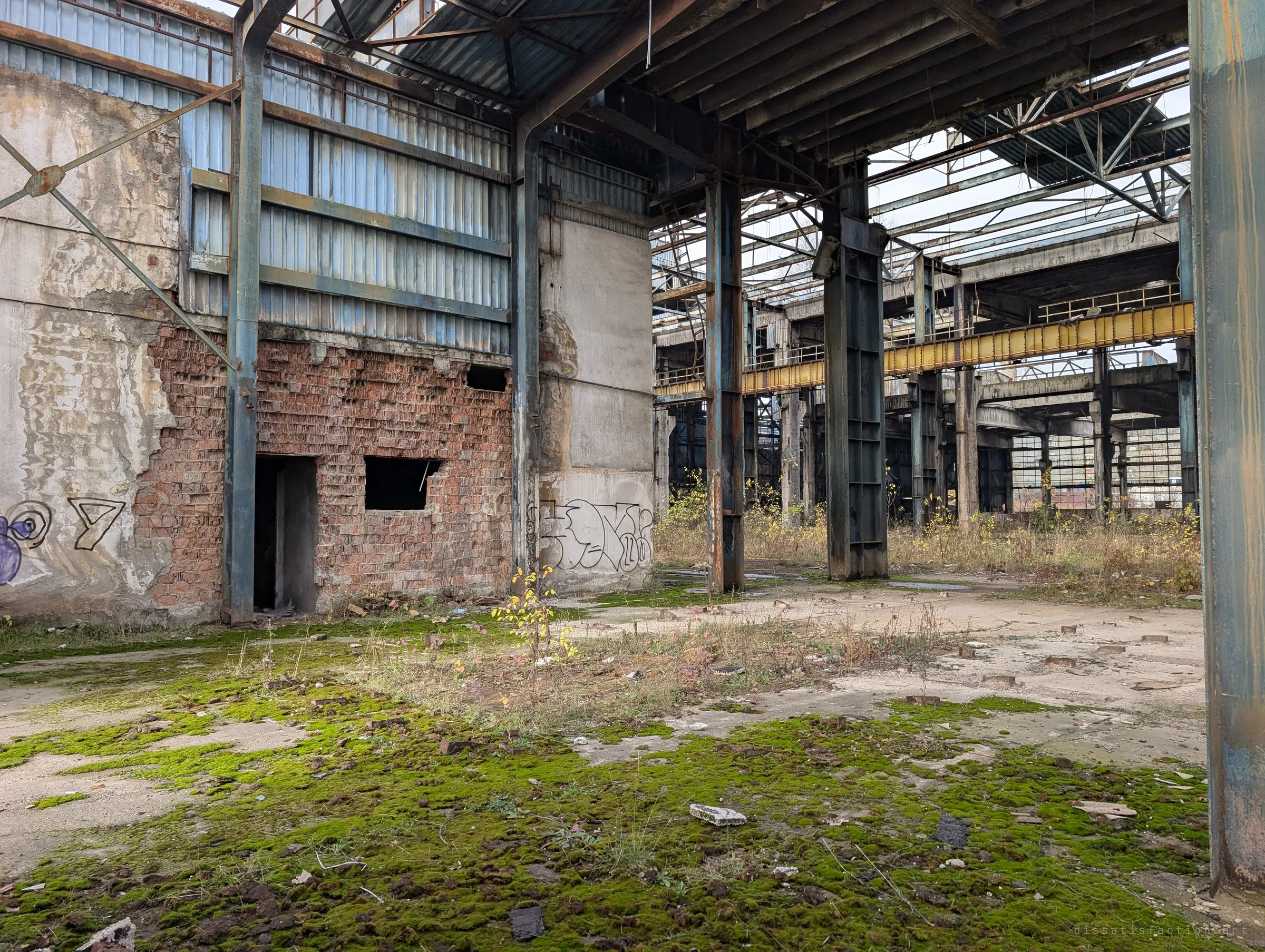 A half-stripped brick and plaster wall rises beside steel columns, with a yellow crane girder crossing the hall beyond. Moss and saplings are reclaiming the floor.