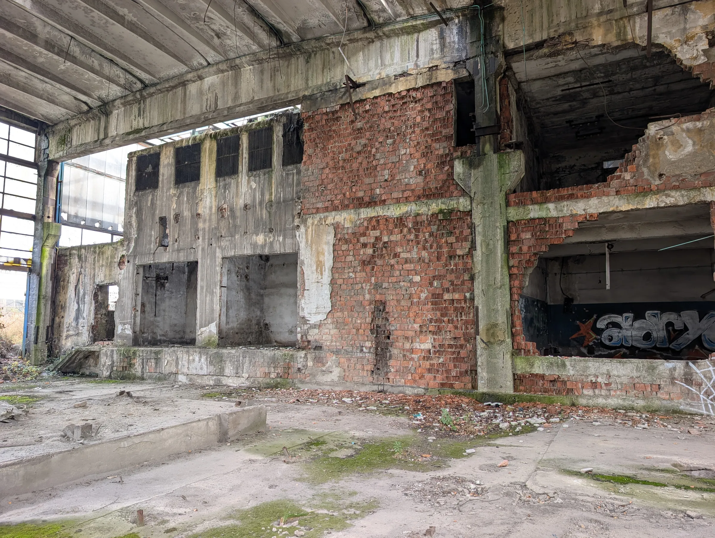 A gutted two-storey block inside the hall, part bare brick and part stained concrete, with a torn-open upper room showing graffiti. Rubble spills across the floor below.