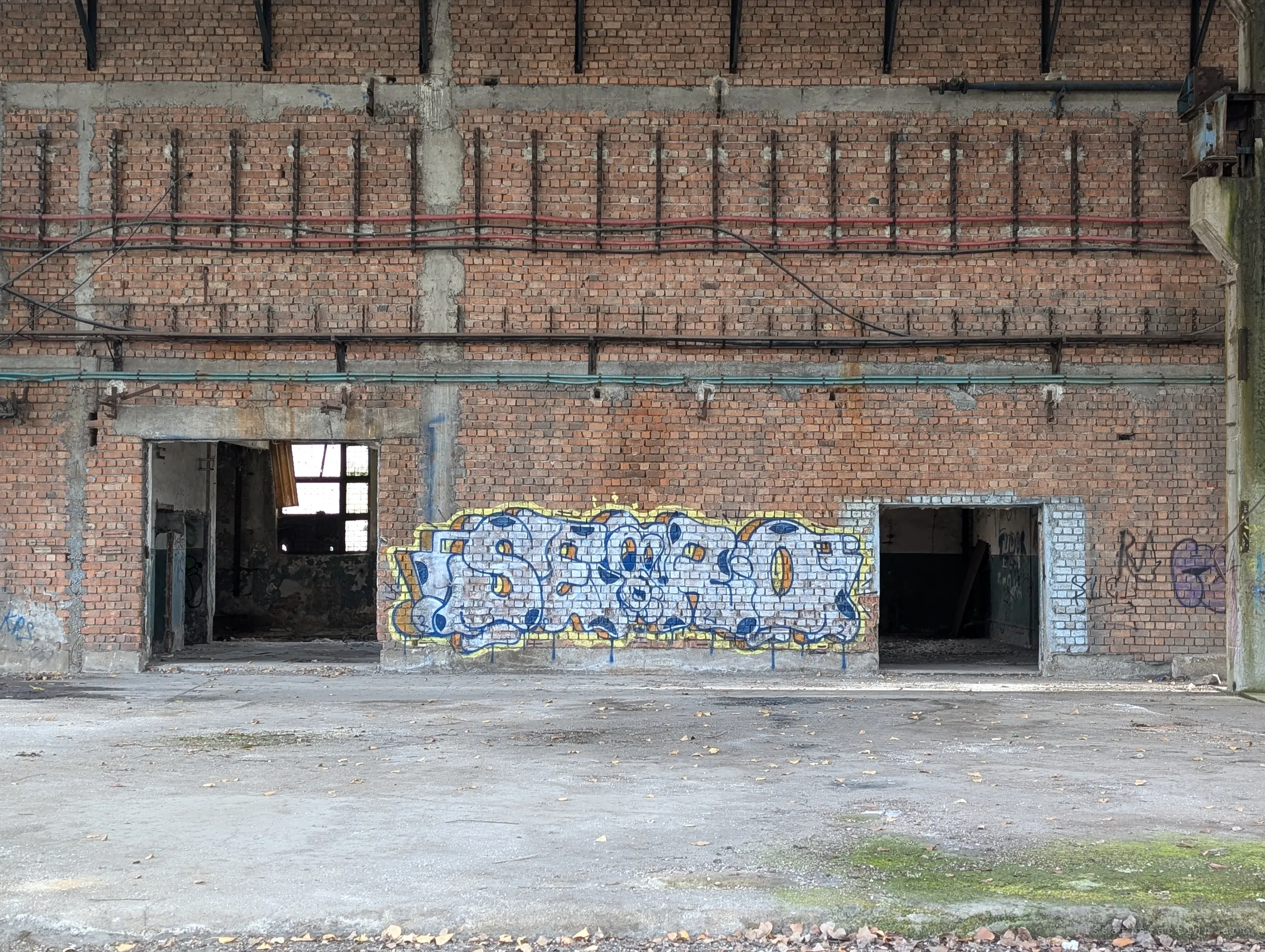 A flat brick wall laced with old cable runs, a bright blue and yellow graffiti piece between two dark doorways. A calm, symmetrical study of the hall interior.