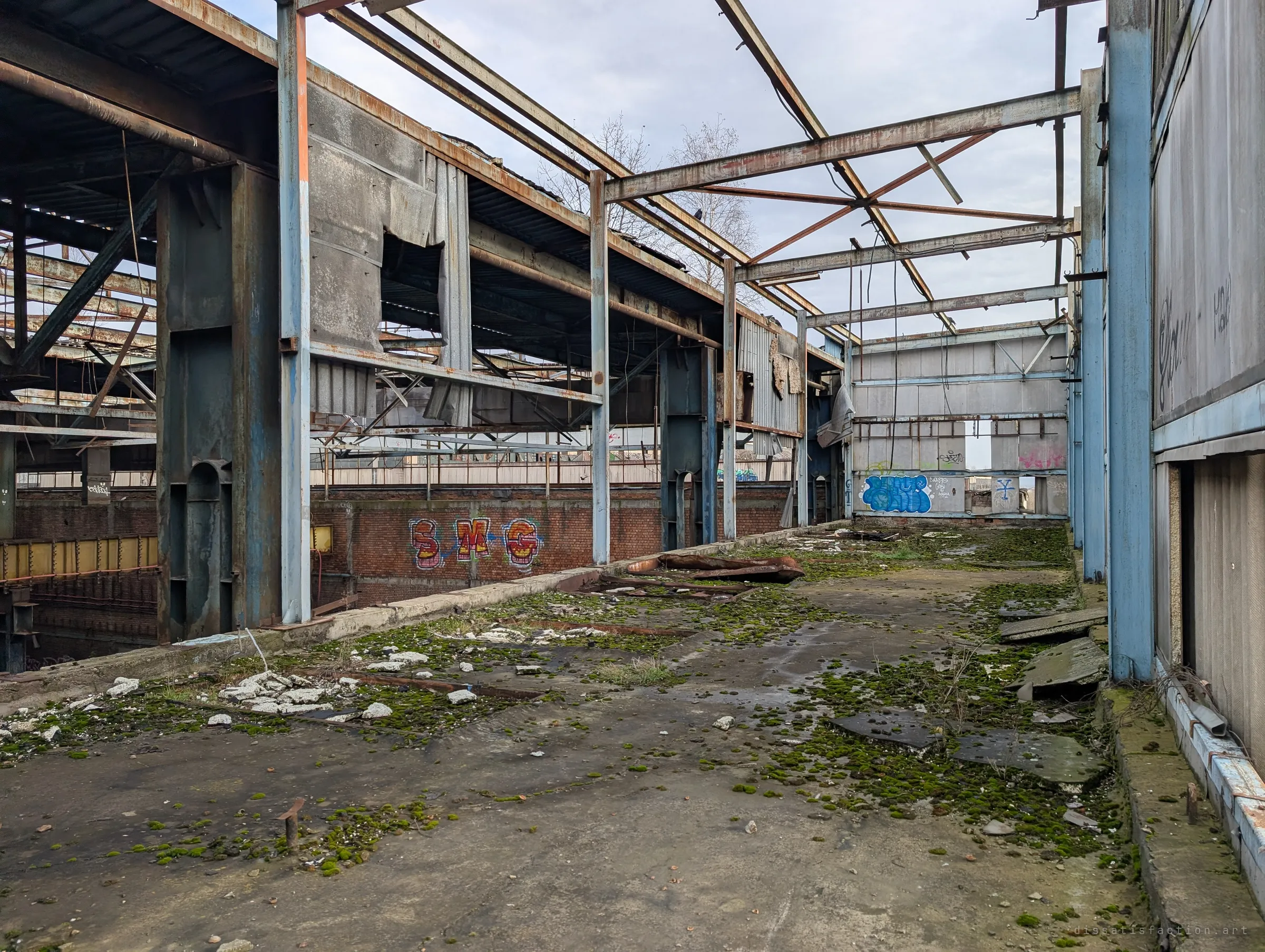 An upper-level walkway covered in moss and fallen roof sheeting runs between rusted steel frames, overlooking graffitied brick below. Shows how far the roof has come apart.