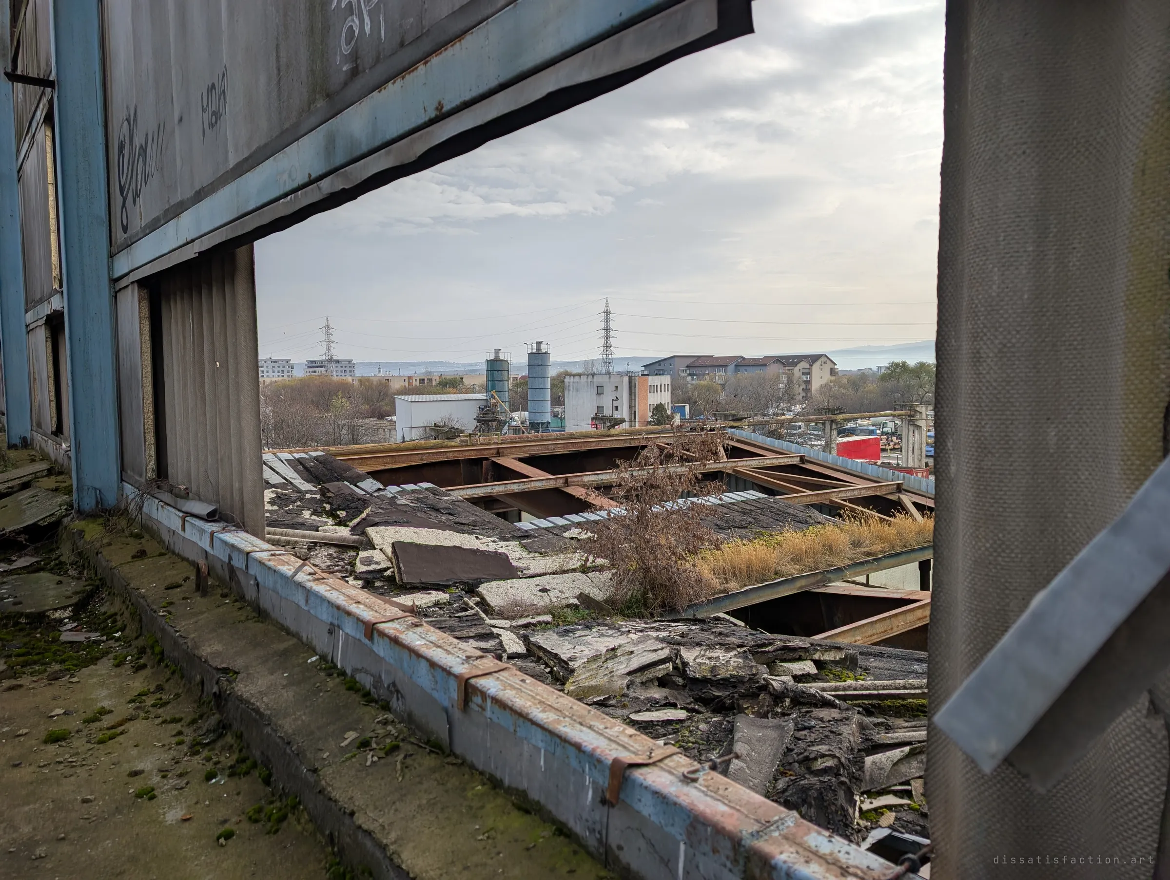 A torn wall panel frames a view over a collapsed, weed-grown roof toward silos, pylons and apartment blocks under a grey sky. The rot inside meets the living city just past the edge.