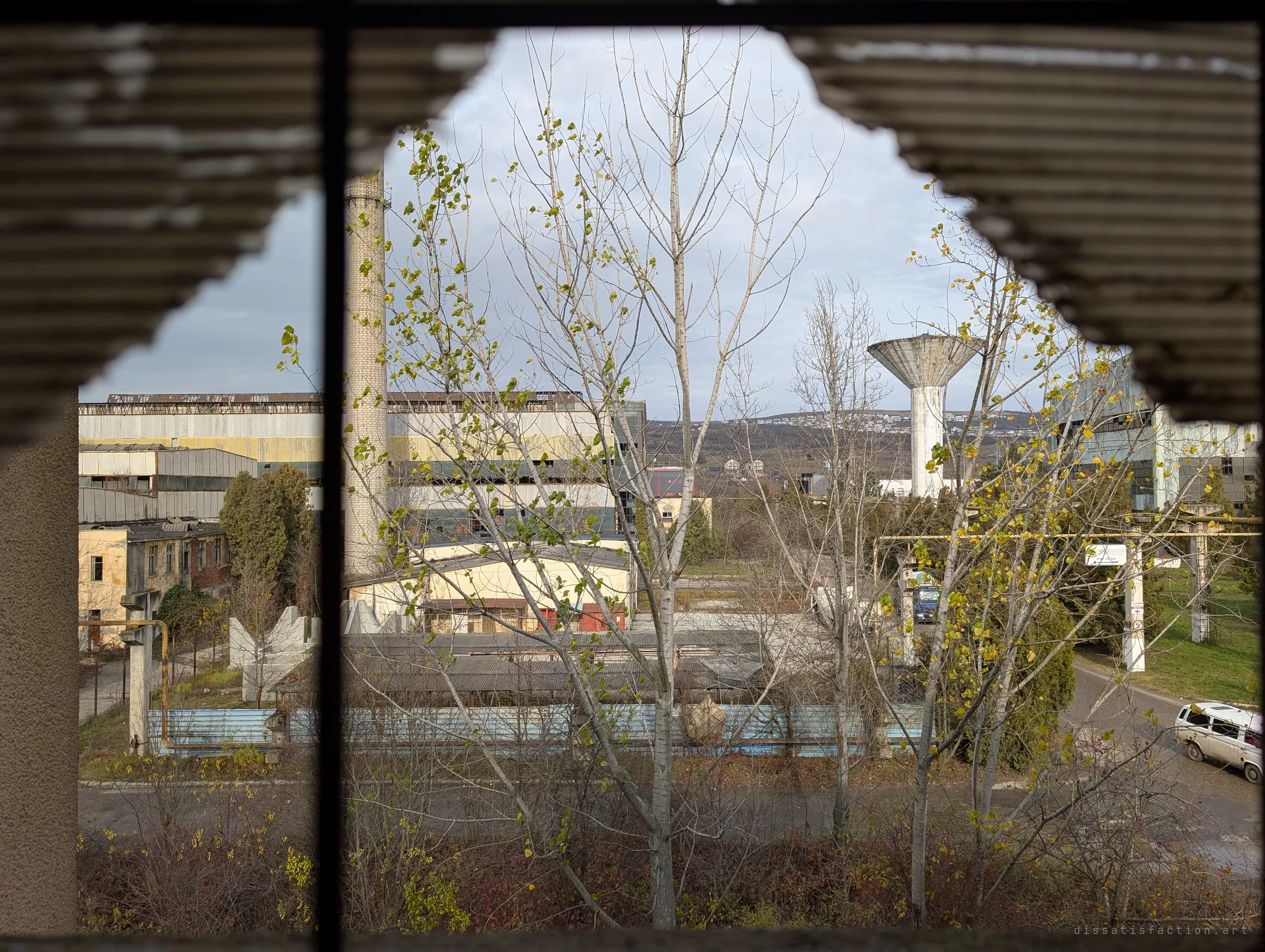 Through a jagged hole in corrugated cladding, the wider works spread out: a brick chimney, a concrete water tower, long factory halls and an access road. The scale of the complex shows in one frame.