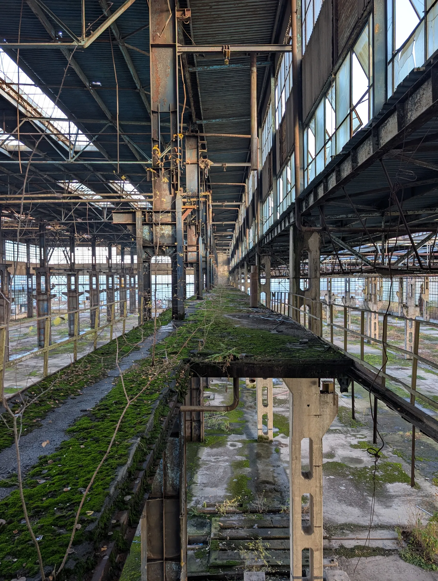 A moss-covered crane rail walkway runs the length of the hall between rows of steel columns and broken clerestory windows. Saplings are growing out of the elevated track.