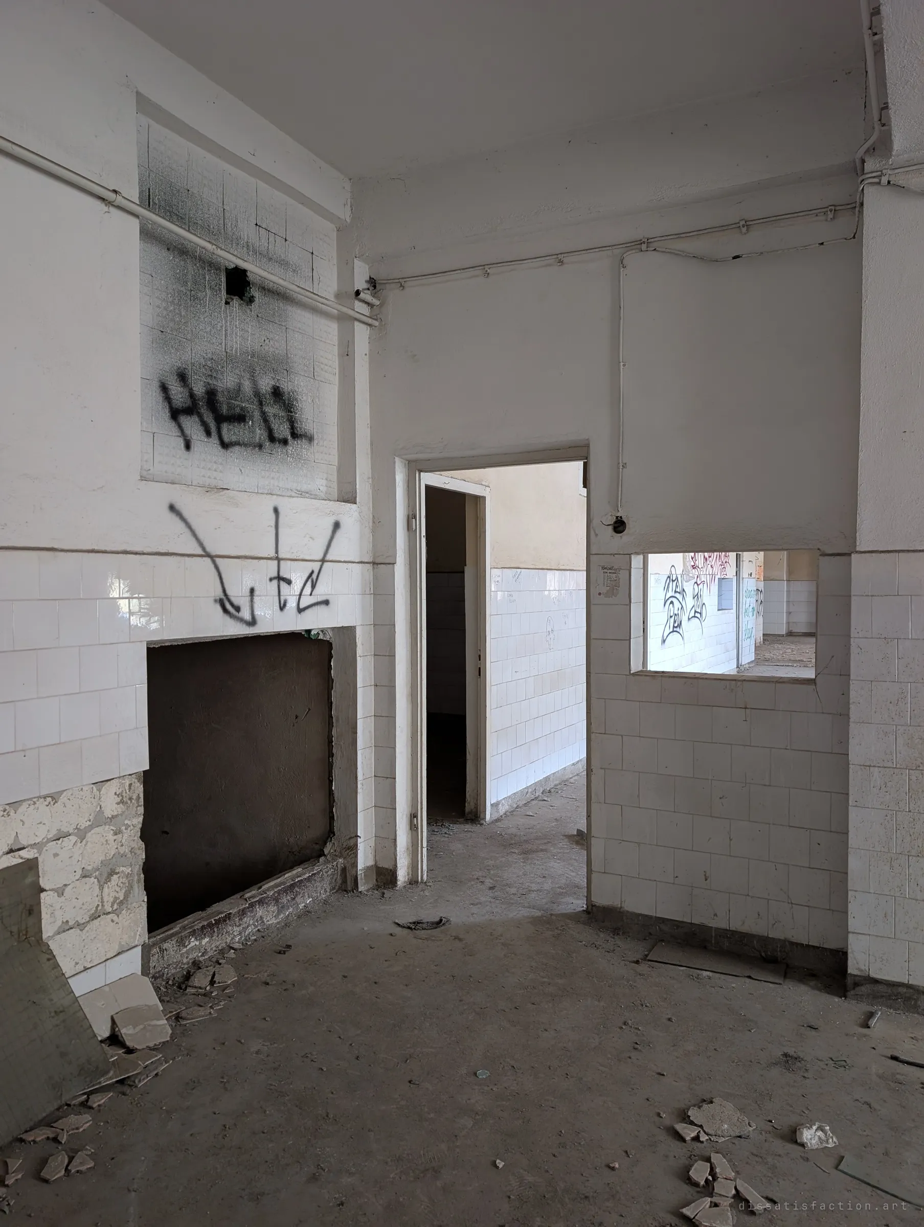 A white-tiled washroom stripped of its fittings, with a gaping hole in the wall and graffiti arrows pointing into it. A doorway and a serving hatch look through to more tiled rooms.