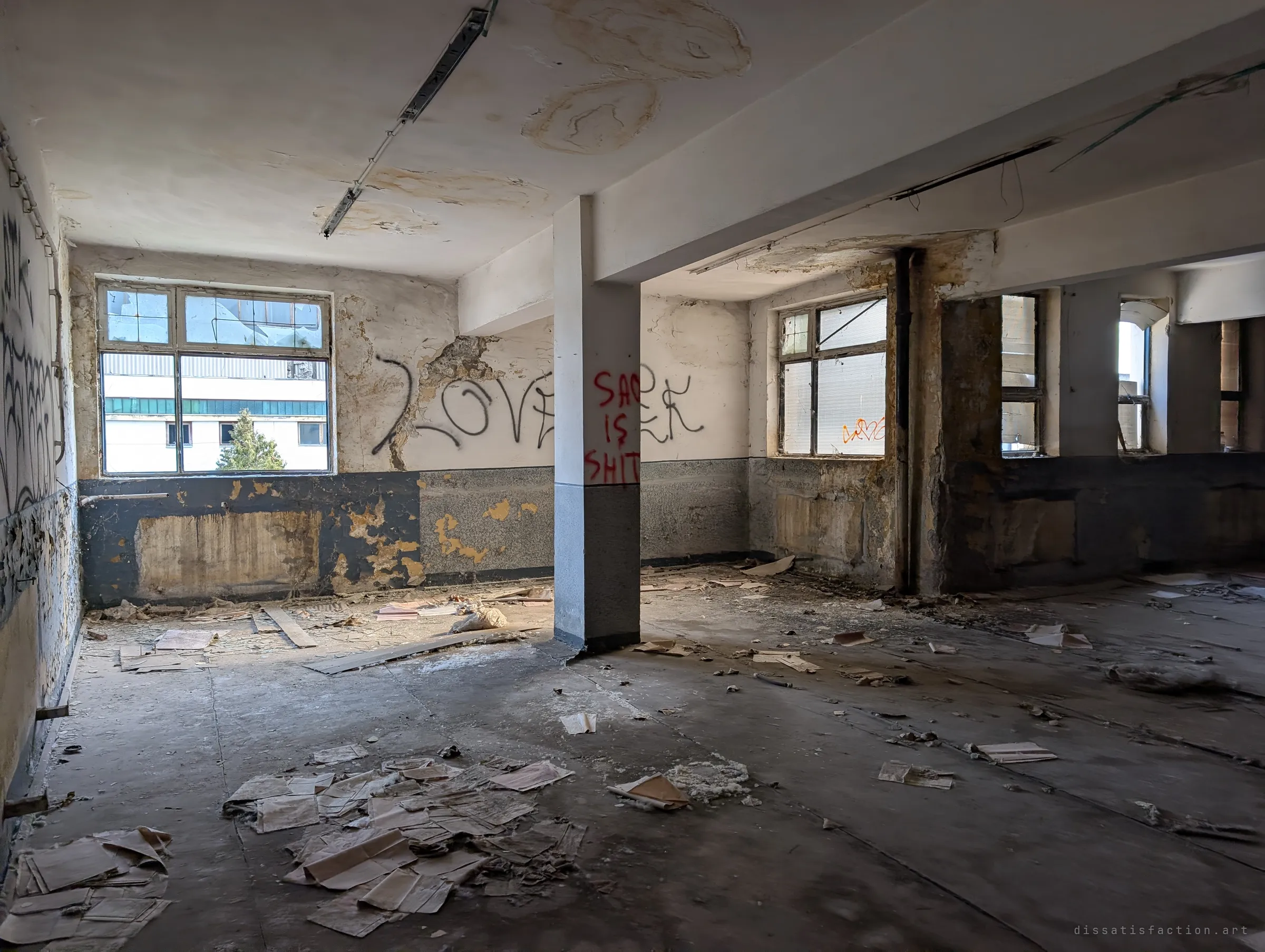 An office floor littered with loose papers and plaster, sprayed love-themed graffiti across the walls and a central pillar. Light from the windows falls across the mess.