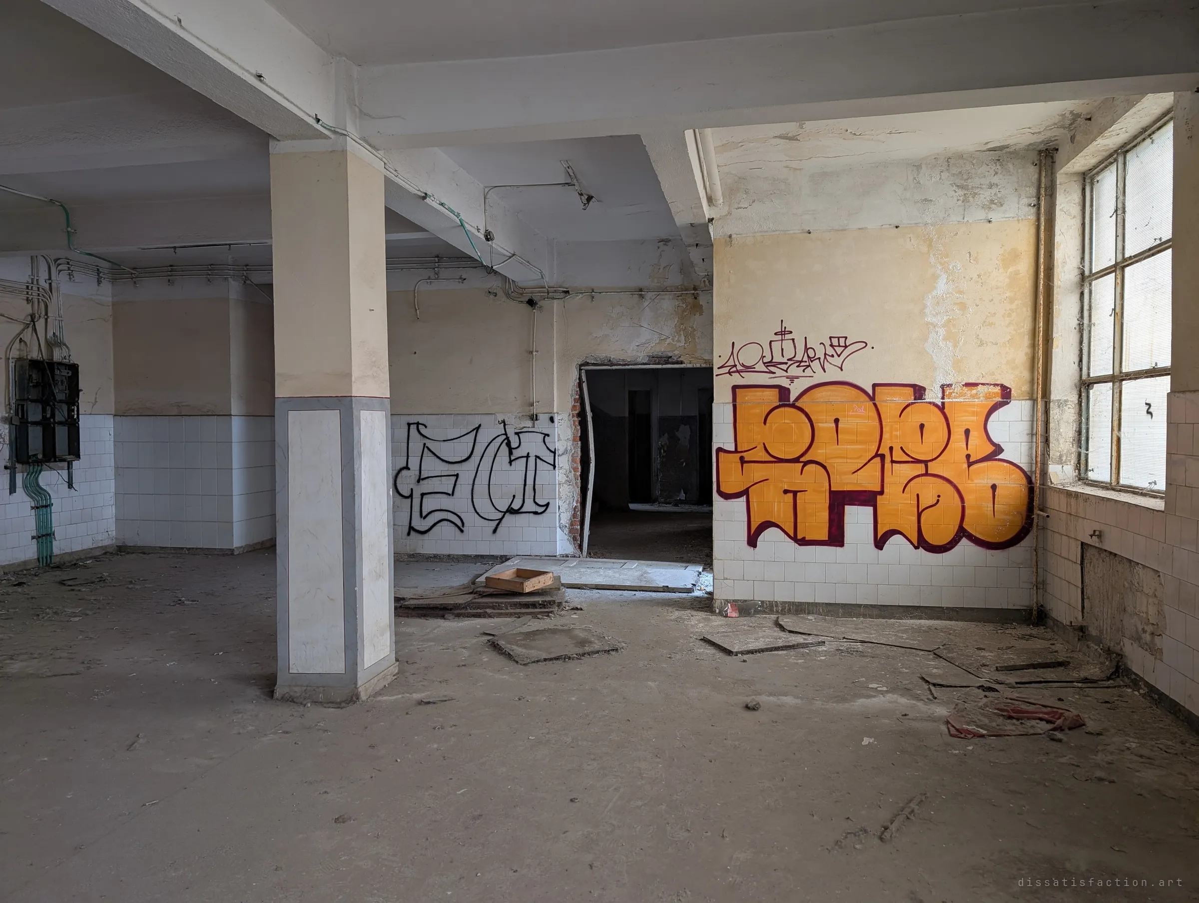 A tiled hall with a marble-clad pillar, an old fuse cabinet on the left and a large orange graffiti piece beside a dark doorway. The bright window light makes the empty room feel almost clean.