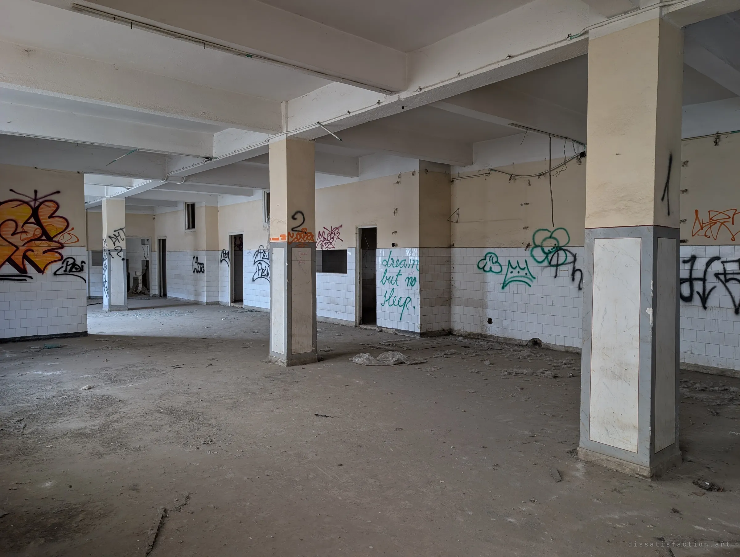 A wide hall of numbered pillars and white tiles, the walls covered in tags and a handwritten line about dreaming without sleep. Doorways lead off into further empty rooms.