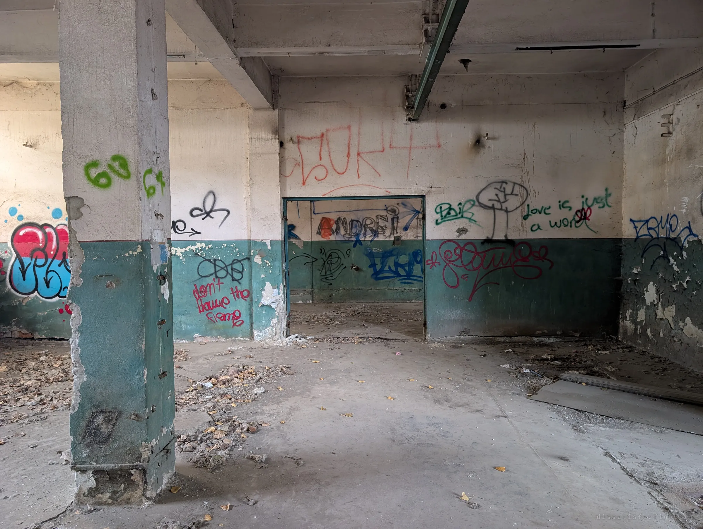 A green-and-white room thick with tags and scrawled lines about love, a doorway opening onto another graffiti-covered space. Dry leaves have blown across the concrete floor.