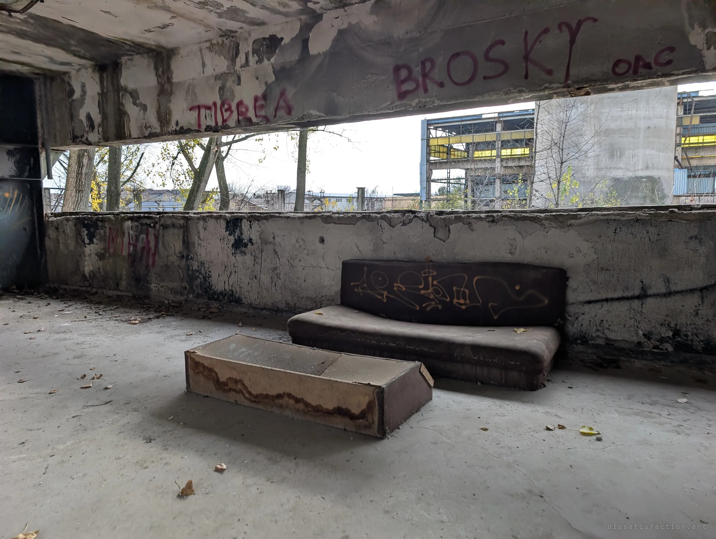 A worn sofa and an overturned drawer sit against a low wall on an open floor, names sprayed on the beam above. Beyond, bare trees and the skeleton of another factory hall.