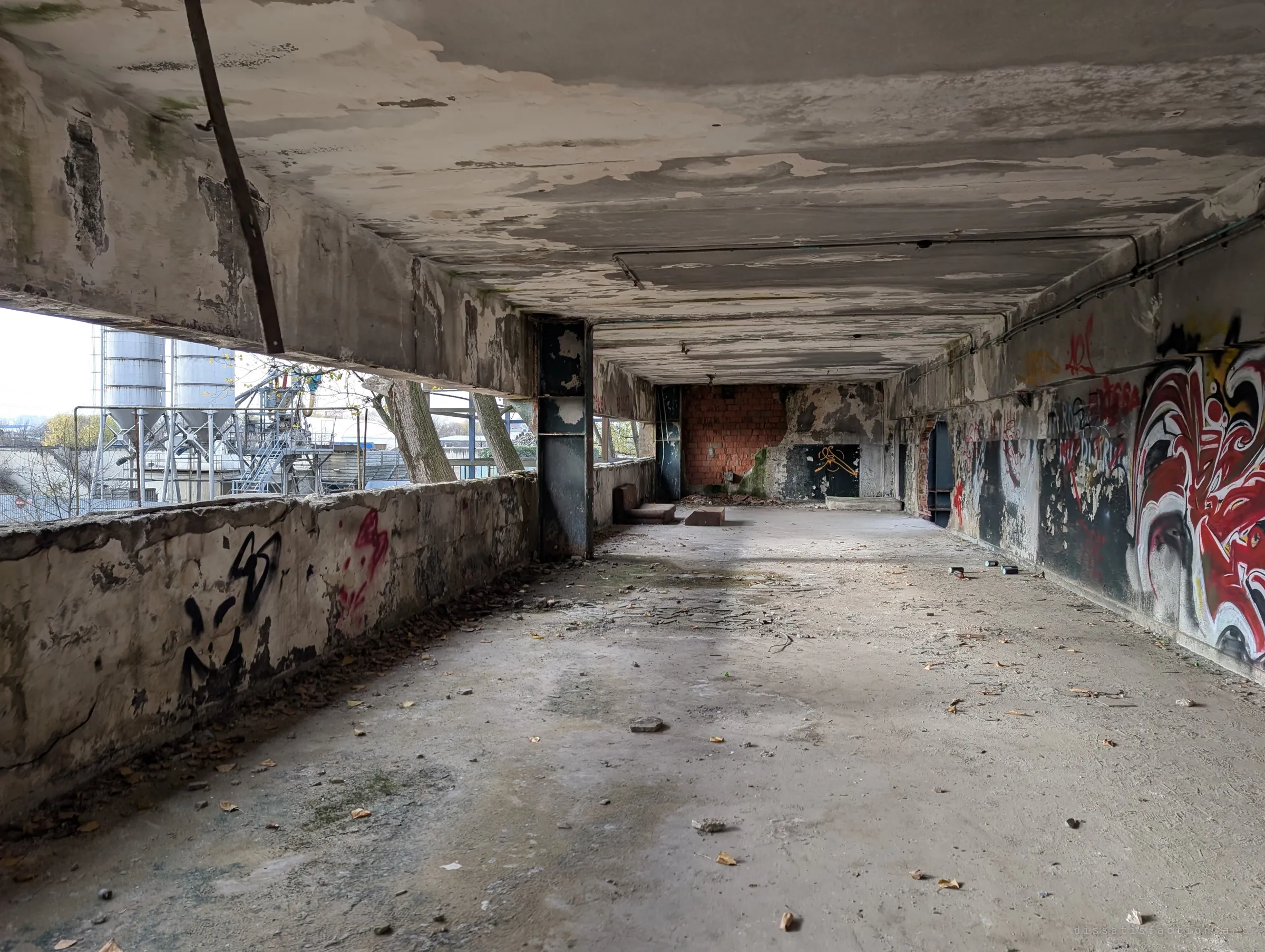 A long open-sided gallery with a stained, peeling ceiling, a red-and-white mural running down one wall and silos visible outside. It reads like a stage left for the painters.
