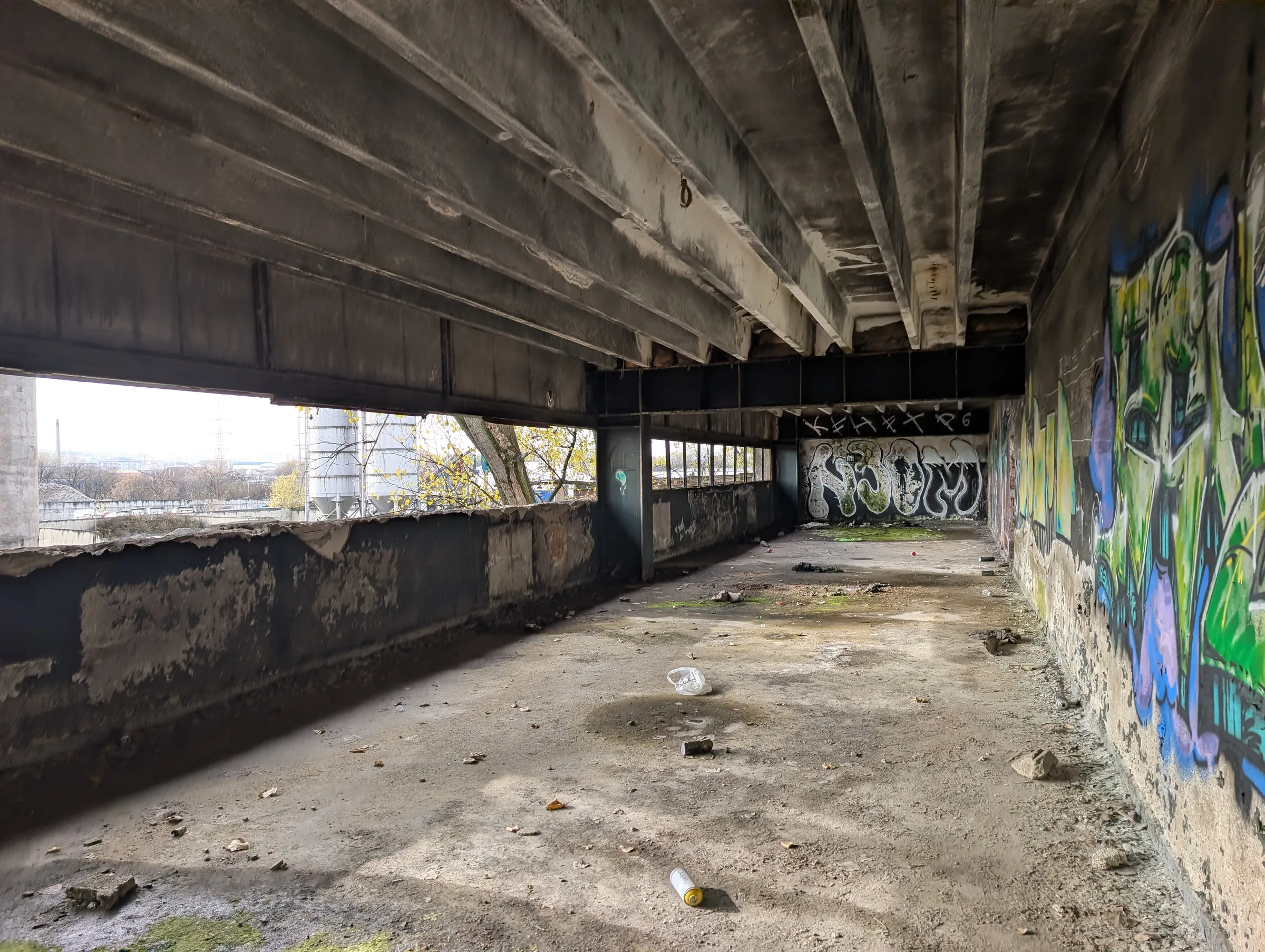 Heavy concrete ribs run over an open gallery, a bright green mural on the right and spray cans scattered on the floor. The far wall carries a big black-and-white piece.